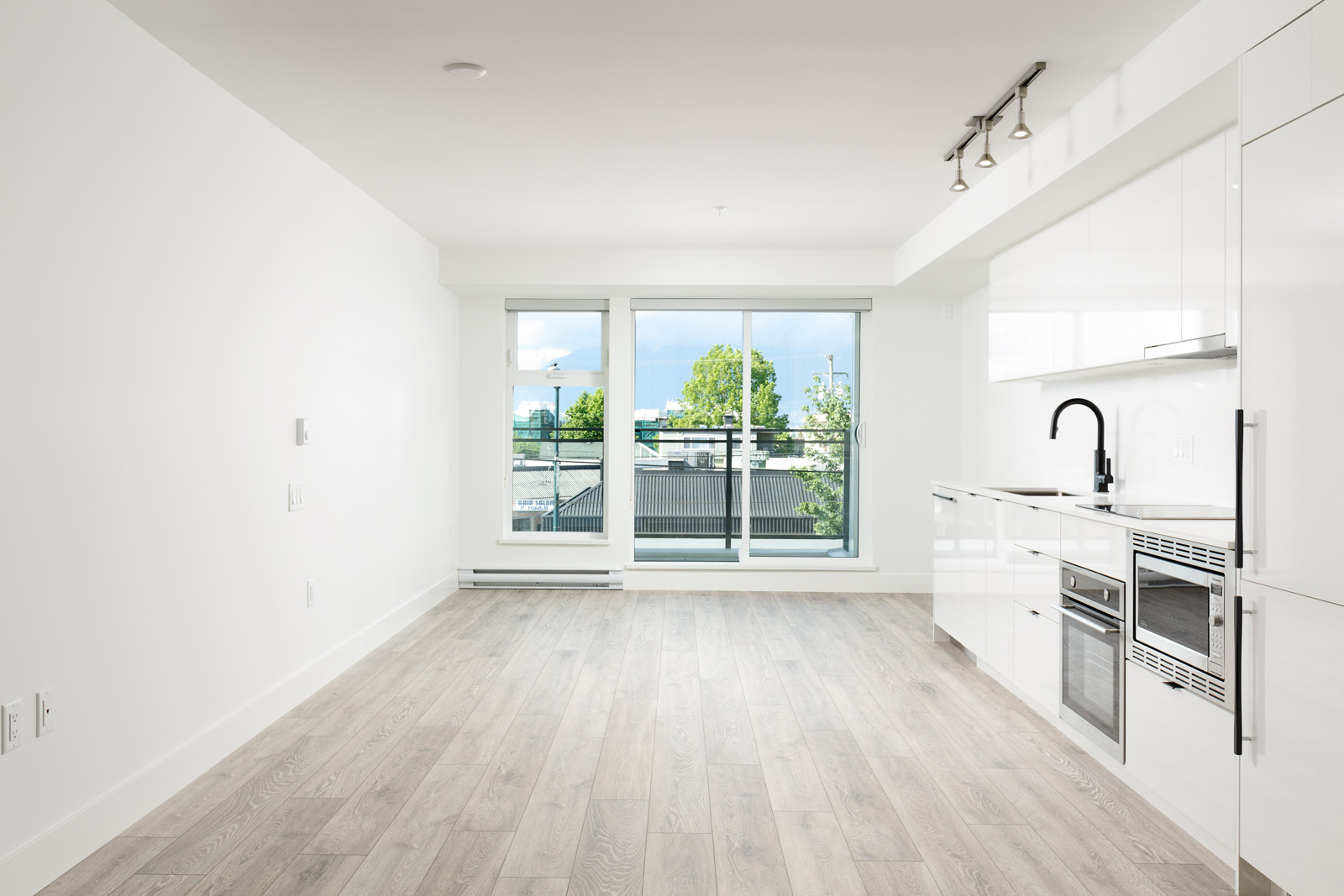 A modern, unfurnished apartment with white walls and light wood flooring, featuring a sleek kitchen area and large windows letting in natural light.