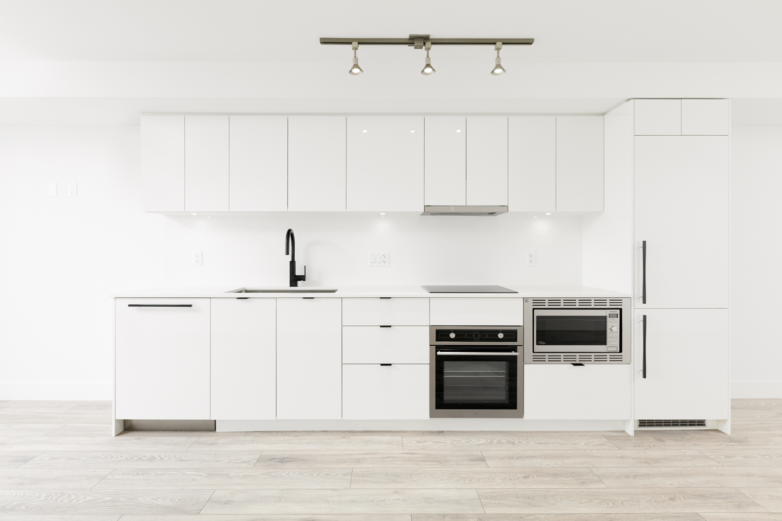 Minimalist white kitchen with sleek cabinetry, built-in appliances including an oven, microwave, and stovetop, a black faucet on the sink, and hardwood flooring under a ceiling light fixture.