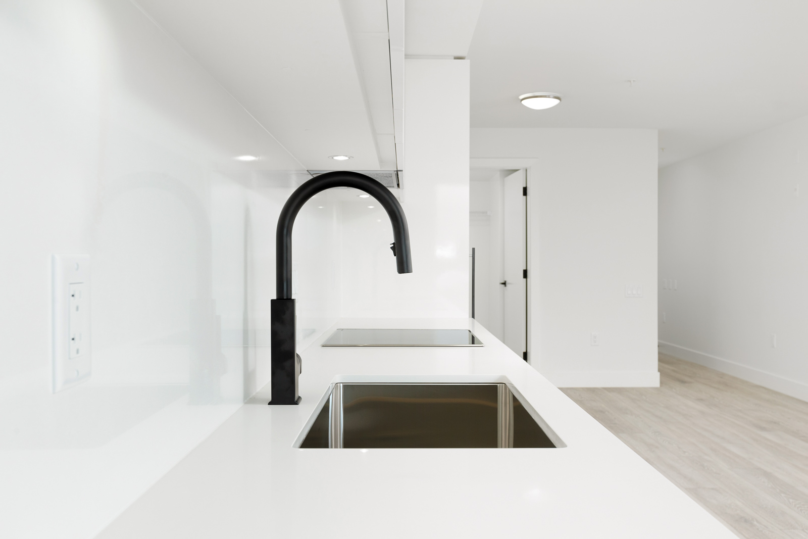 Modern kitchen with a minimalist design featuring white countertops, a black faucet, an electric stovetop, and light wood flooring.