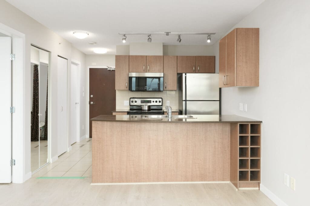 Modern kitchen with light wooden cabinetry, stainless steel appliances, and a beige countertop. There is a wine rack on the side and track lighting on the ceiling. The entrance door is visible in the background.