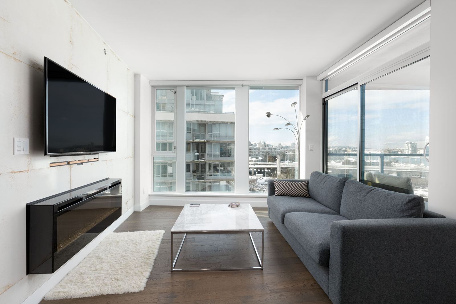 condo living room with floor to ceiling windows with flat screen tv on left wall and sofa on right and coffee table in centre with hardwood floors