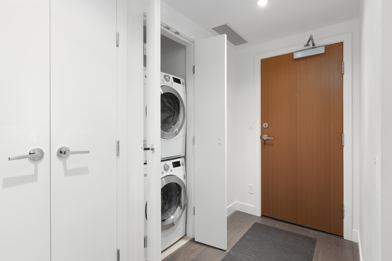 partial view of stacked white washer and dryer with white door on the left and darker wood entrance door to condo unit on the right
