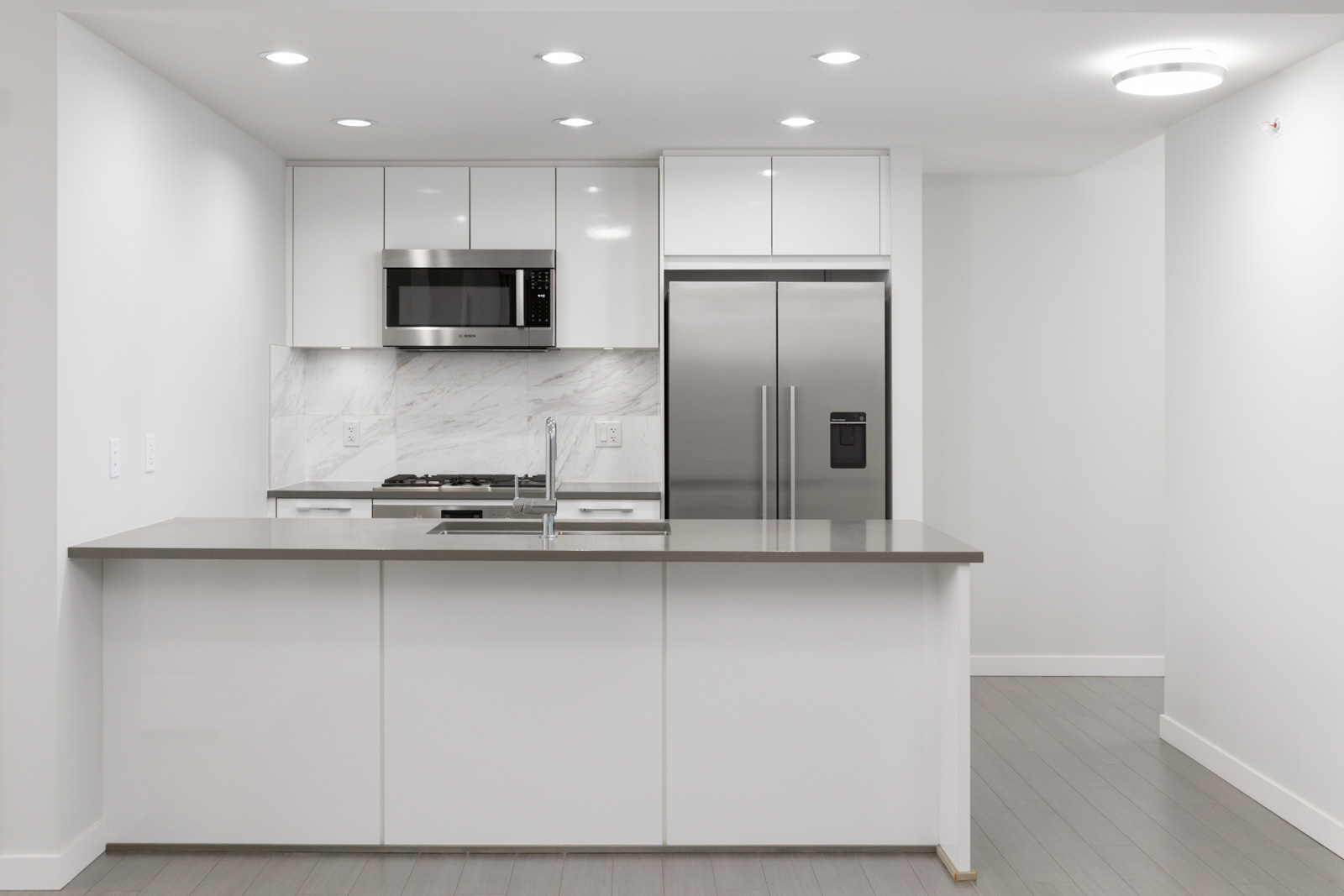 front view of kitchen in condo with with island in front and kitchen wall in back with stainless steel refrigerator on right and over the top microwave on left amid white cabinets