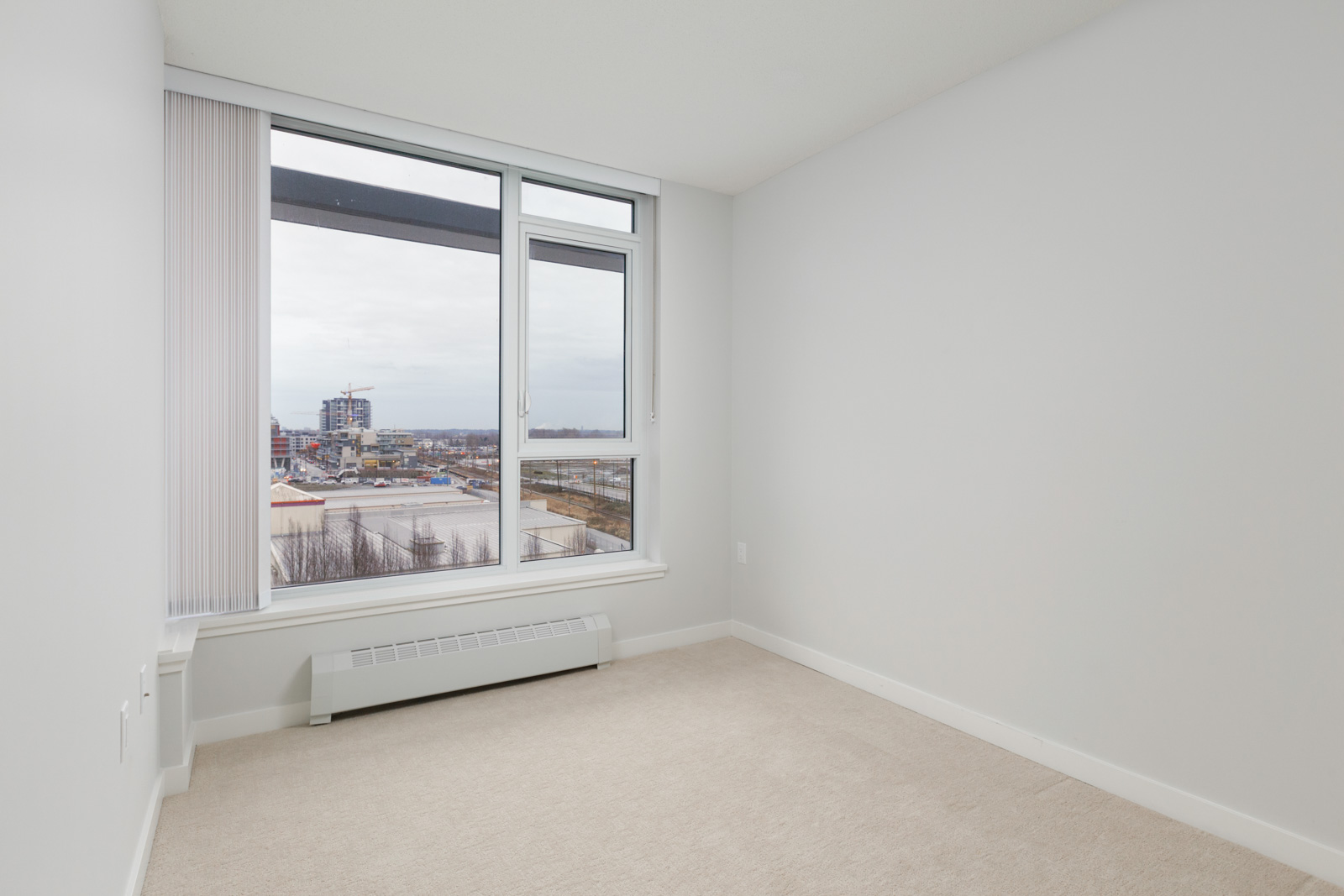 diagonal view of empty condo bedroom with windows on left and white wall on right and carpeted flooring