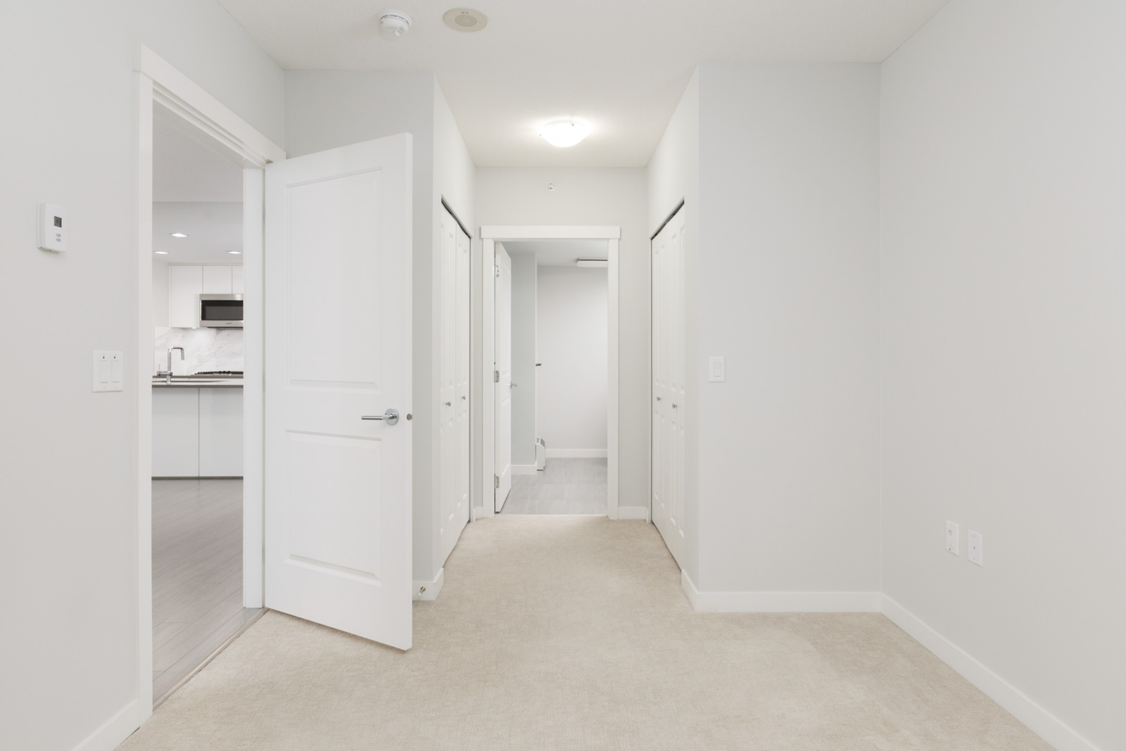 view in long condo hallway with open door into kitchen on left and down hallway in the middle to other rooms with closet doors on both sides of hallway where floors are carpeted and doors and walls are white