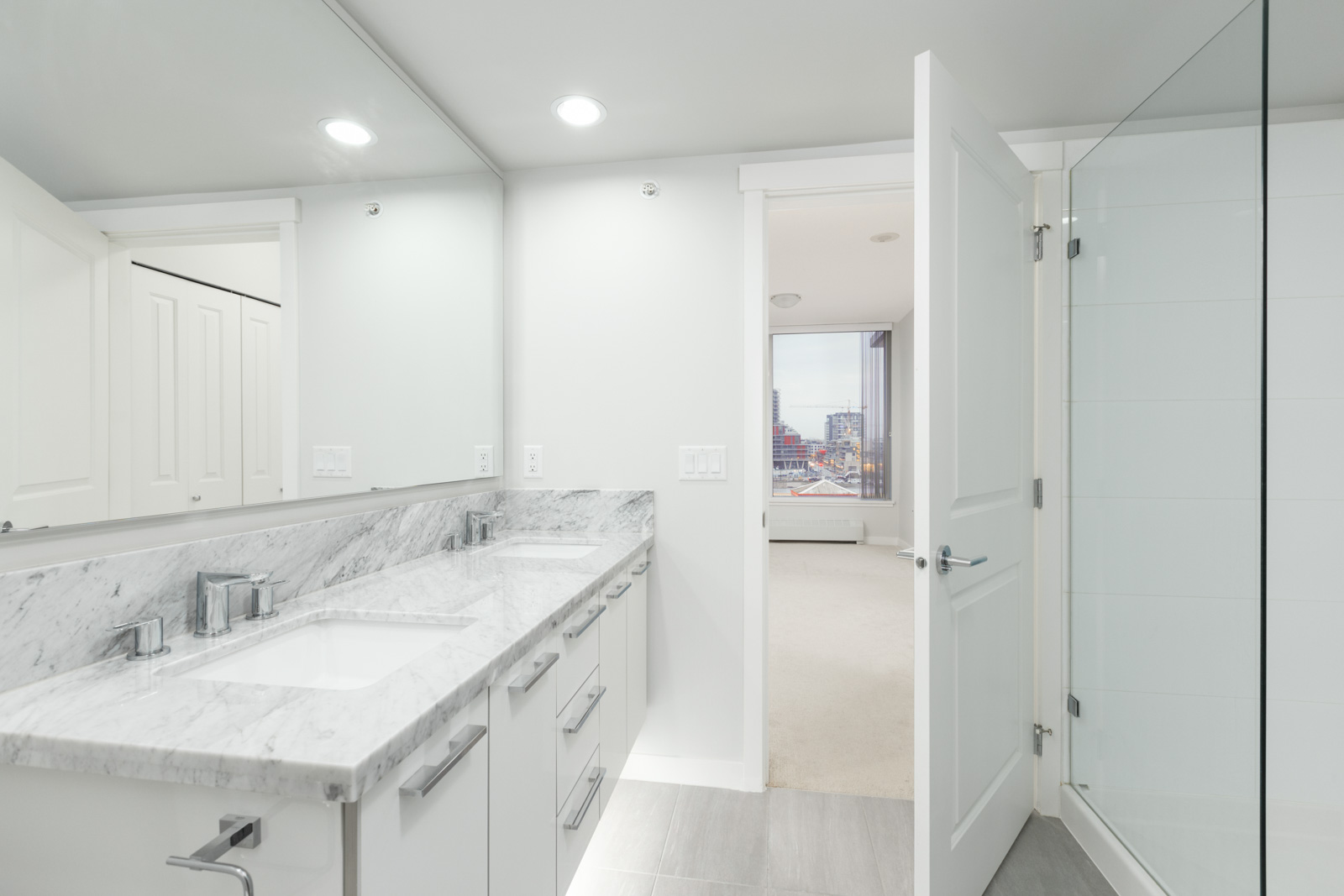bathroom side view with long counter with two sinks and mirror on left and open door to living room on the right with windows to outside in the background