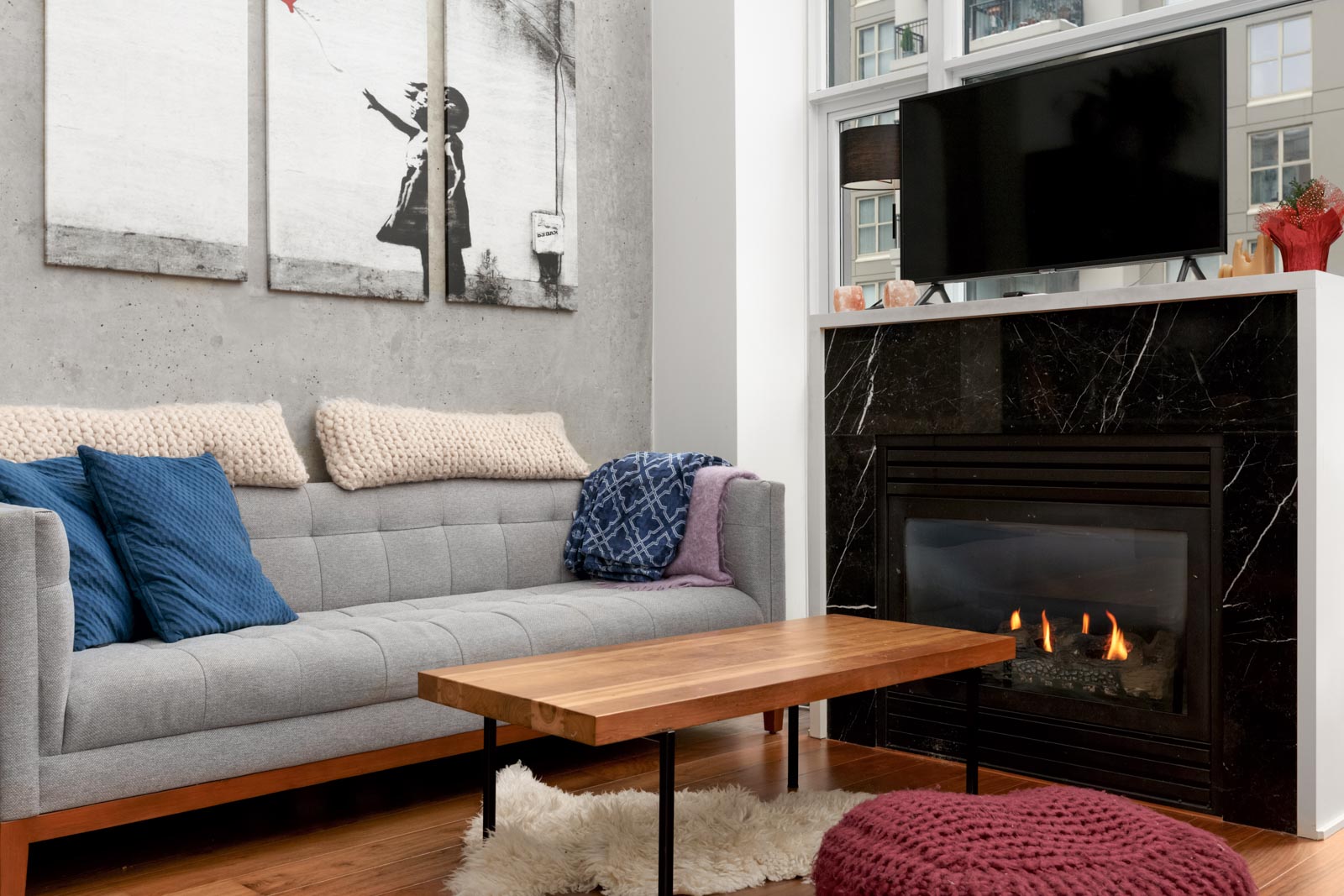 living room of condo with grey sofa on on left with beige throw pillows at the top and blue pillow on the couch along with painting above and coffee table in middle and tv on the right