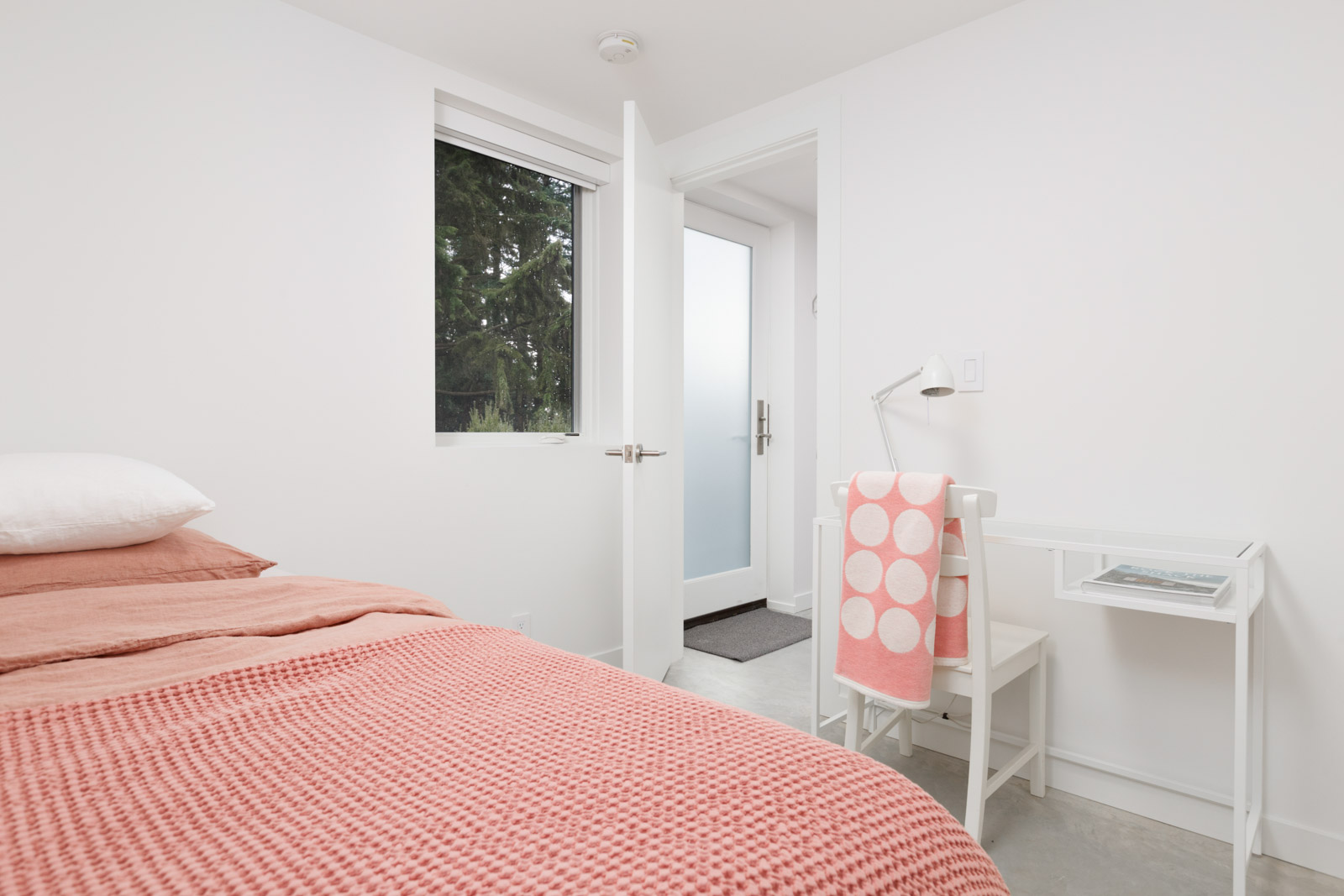 bedroom with rose linens on left and small window in middle with view of backyard trees and open door to hallway