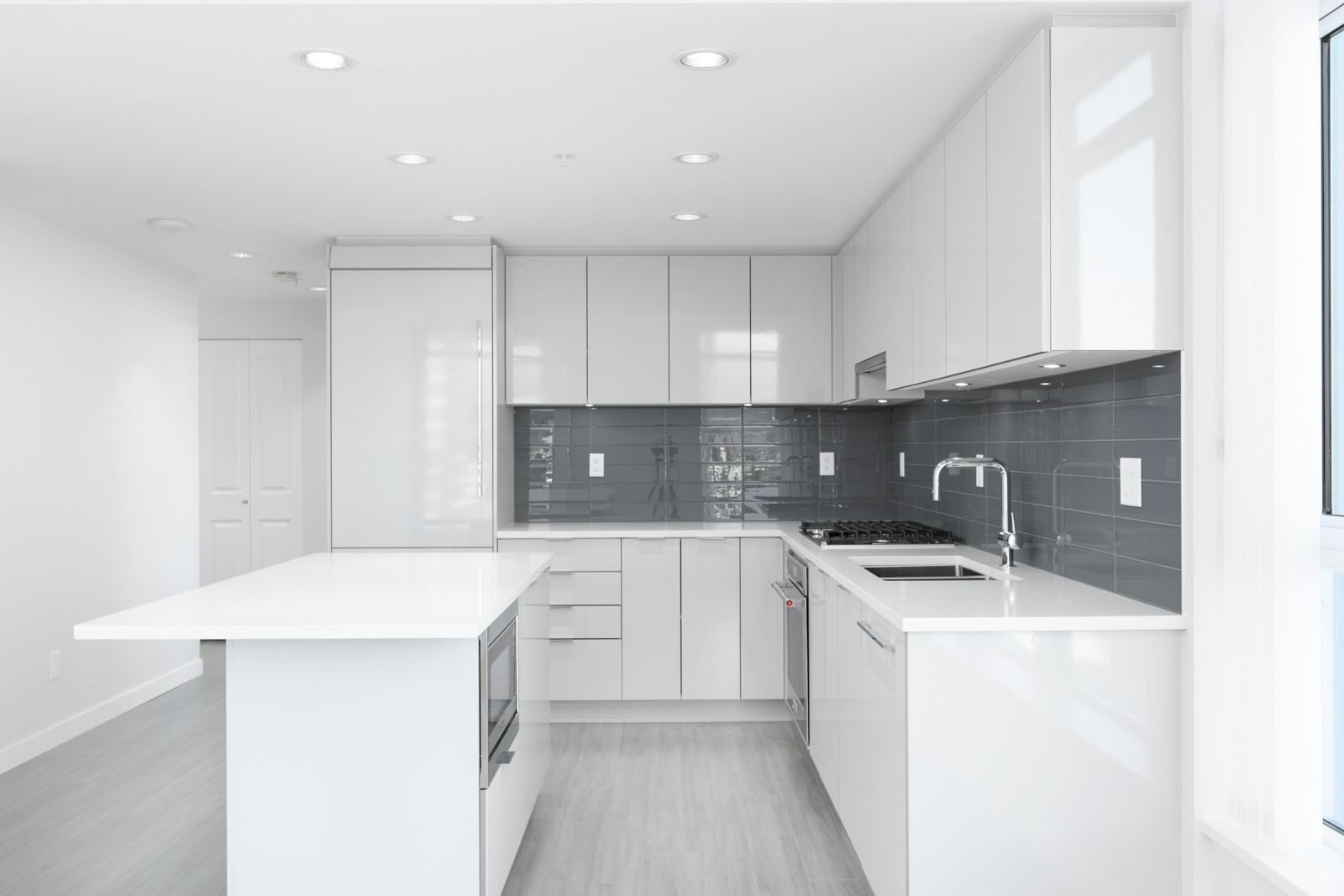 Modern kitchen with white cabinets, gray backsplash, built-in appliances, center island, and light wood flooring, illuminated by recessed ceiling lights and natural light from a window.