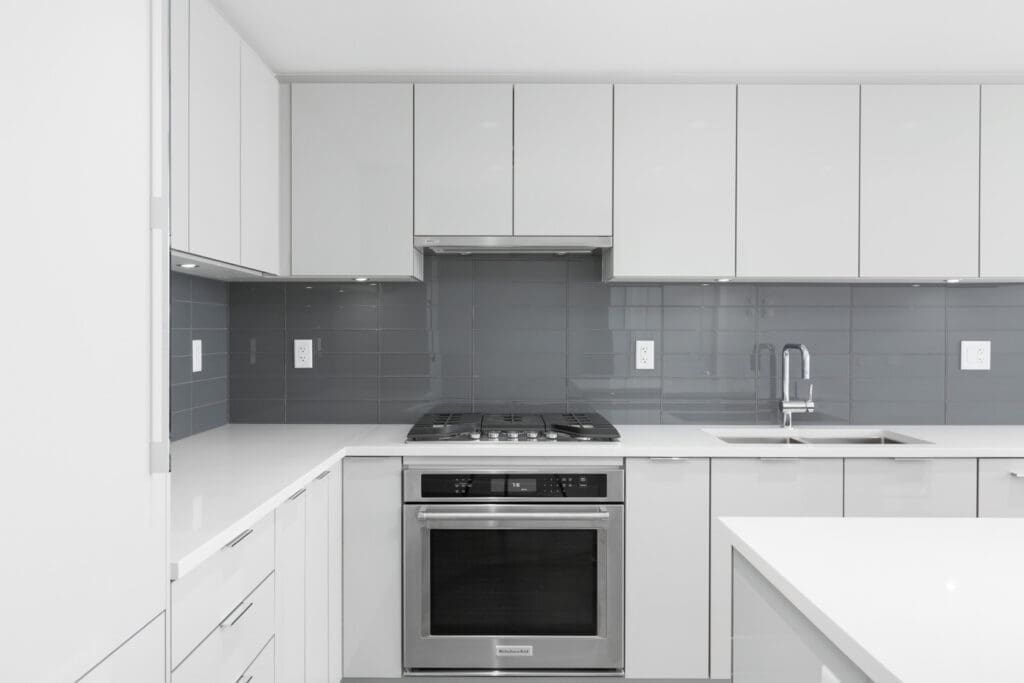 Modern kitchen with white cabinets, stainless steel oven, gas stove, gray tile backsplash, and a sink on the right side. Clean and minimalistic design.