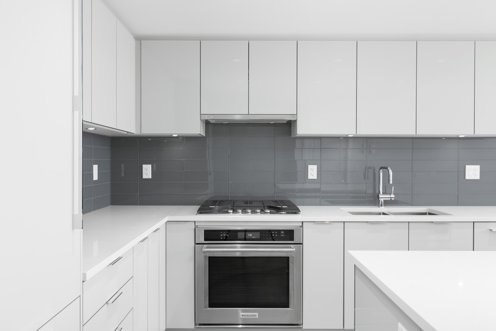 Modern kitchen with white cabinets, stainless steel oven, gas stove, gray tile backsplash, and a sink on the right side. Clean and minimalistic design.
