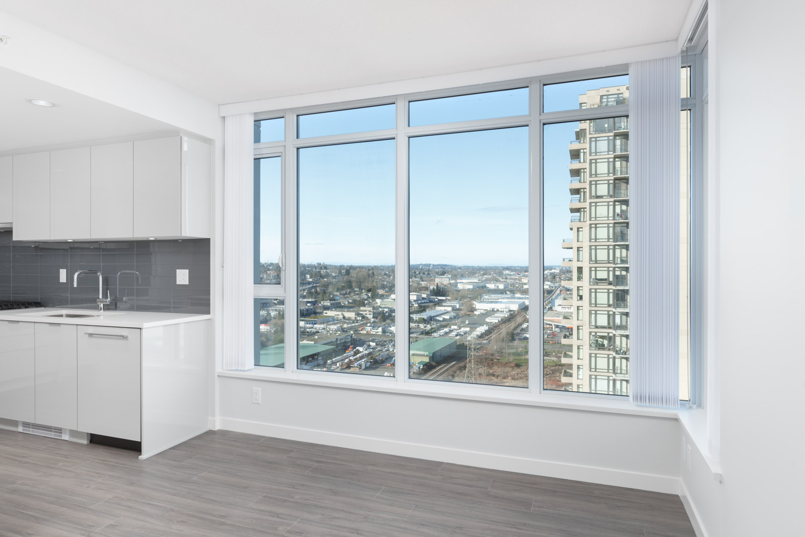 A modern, unfurnished apartment with large floor-to-ceiling windows, gray wood flooring, and a view of a cityscape and distant landscape.