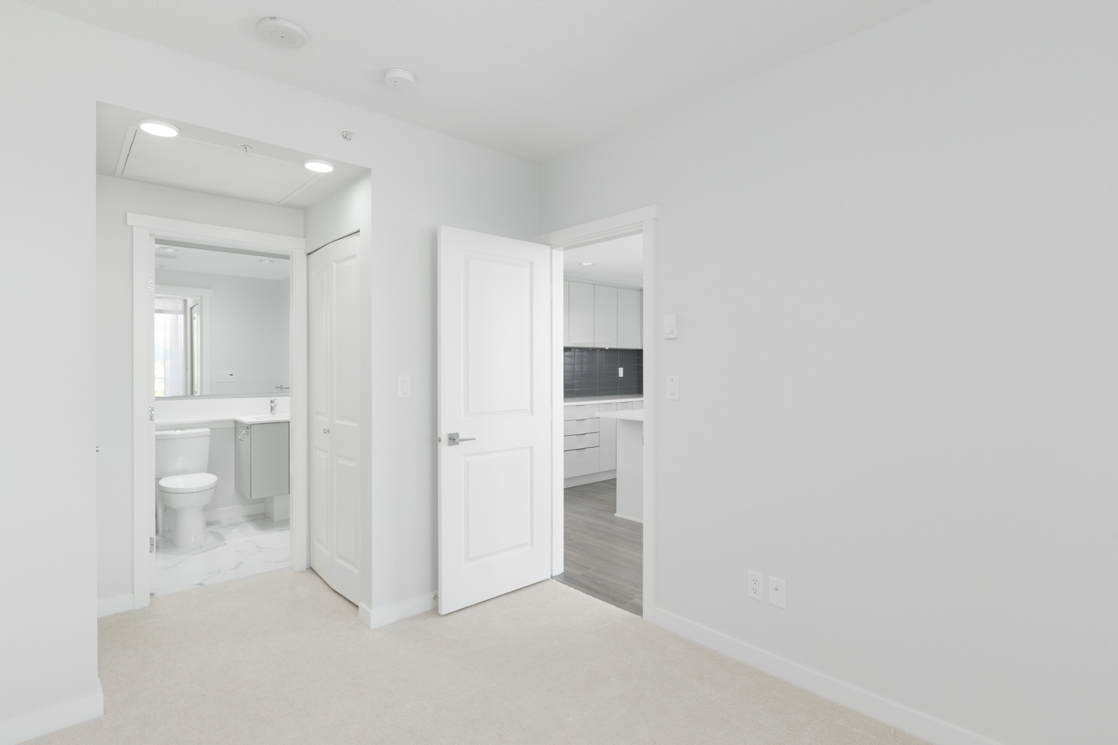 A white-walled, empty room with beige carpet, open doorways leading to a bathroom with a toilet and to a kitchen with white cabinets.
