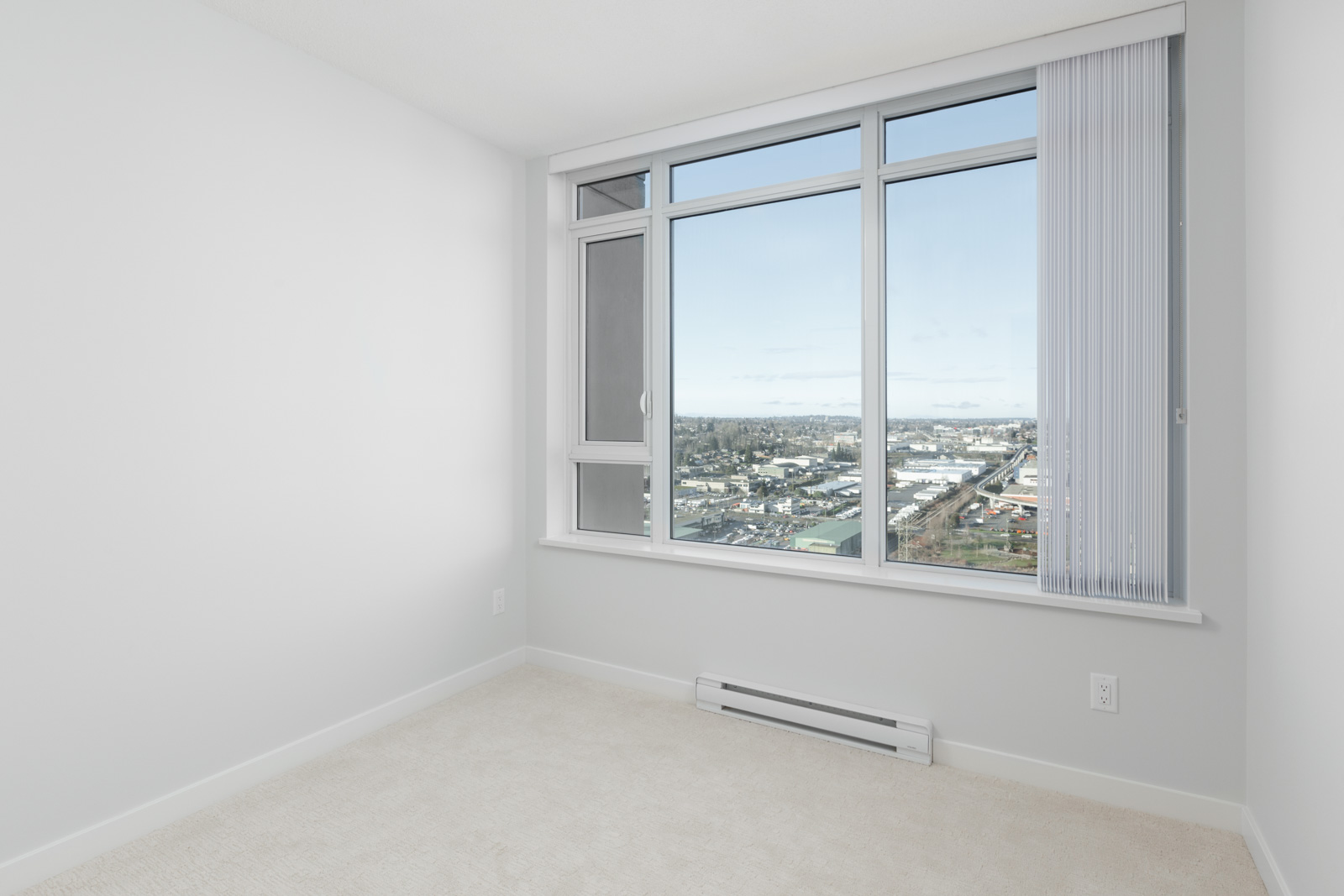 Empty, carpeted room with white walls and large window offering a cityscape view; vertical blinds partially open and natural light filling the space.
