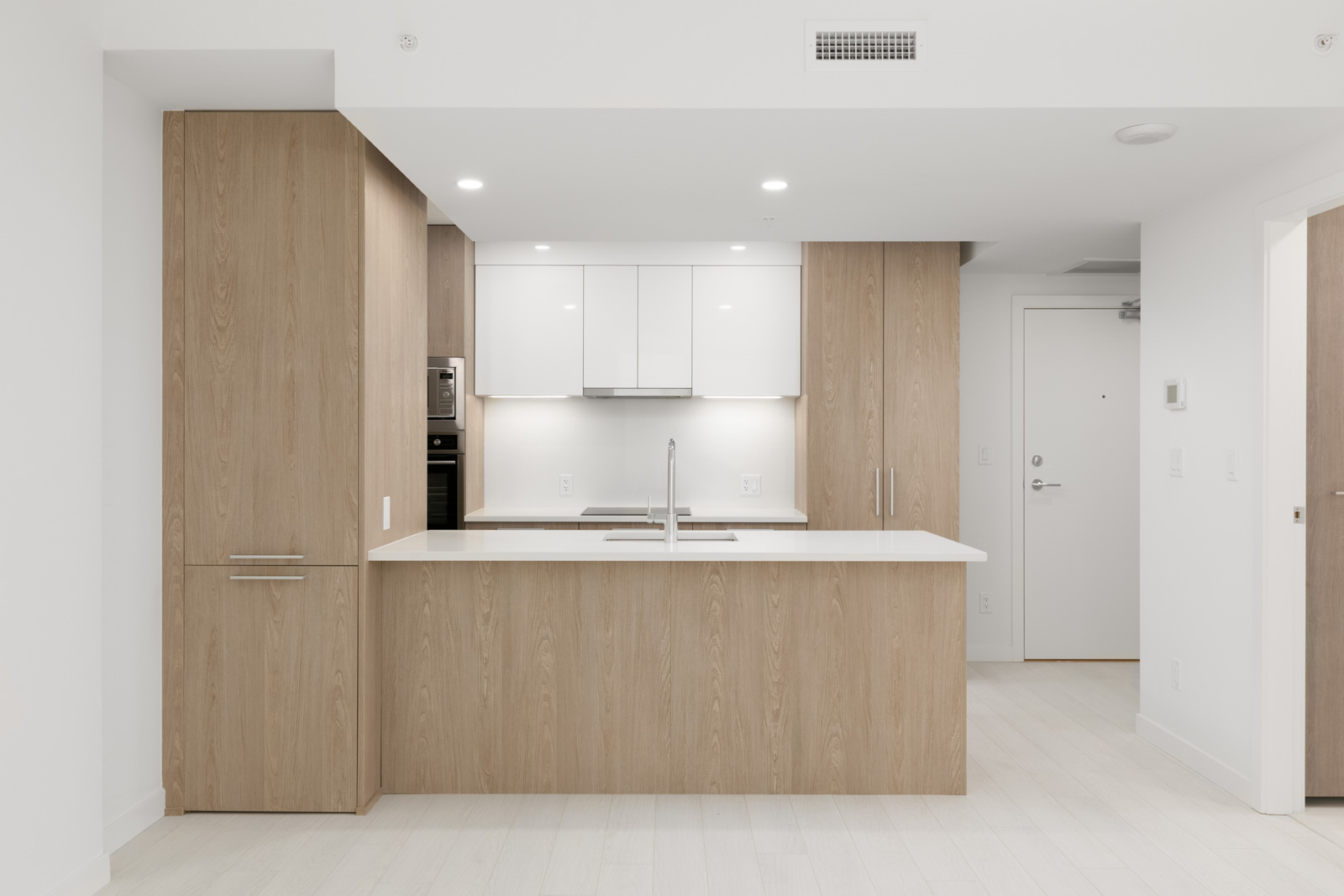 Minimalist kitchen with light wood cabinets, white countertops, and integrated appliances, including an oven and a fridge. The room is bright with modern lighting and white walls.