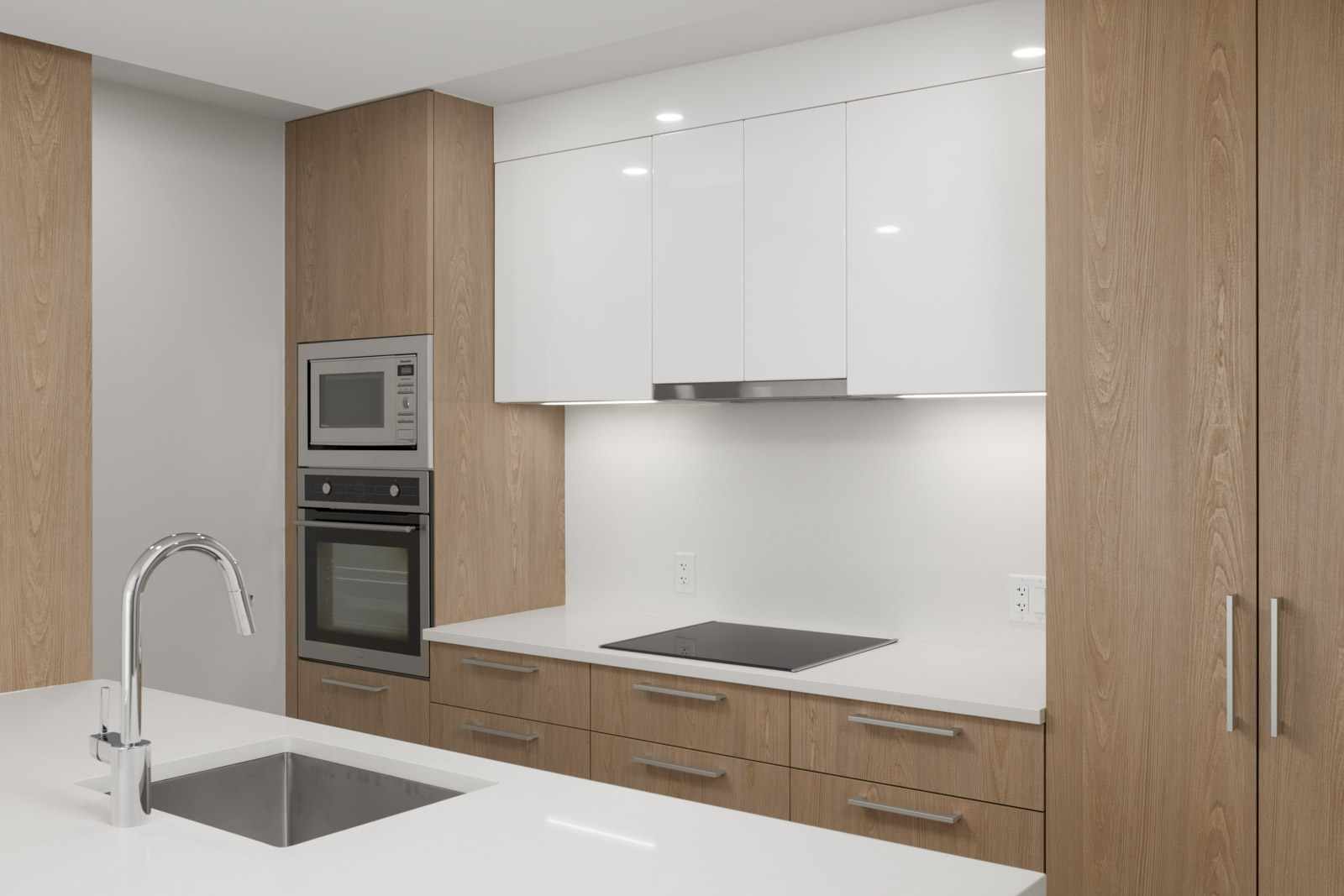 Modern kitchen with wood cabinetry, white countertops, built-in oven, microwave, and electric stovetop. Island in foreground with stainless steel sink and faucet.
