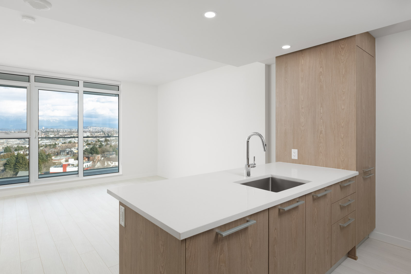 Modern kitchen with wood cabinets and a white countertop, featuring a sink and faucet. The open space includes large windows with a view of a cityscape and mountains in the background.