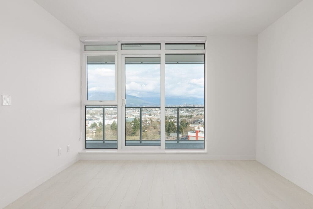 Empty room with light wood floor and large windows. View of cityscape, mountains, and cloudy sky outside.
