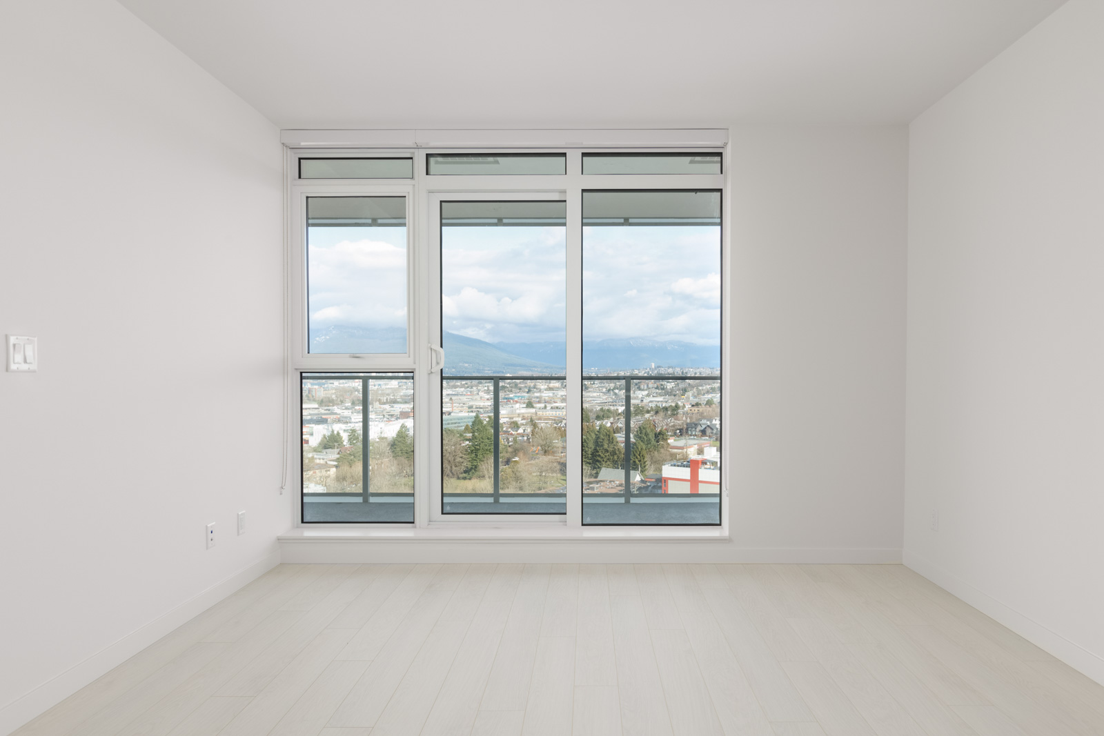 Empty room with light wood floor and large windows. View of cityscape, mountains, and cloudy sky outside.
