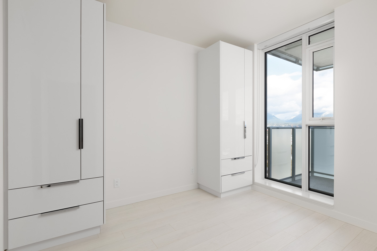 Bright, minimalist bedroom with white walls, light wood flooring, sleek white wardrobes, and a large window door leading to a balcony with a view of hills and cloudy sky.