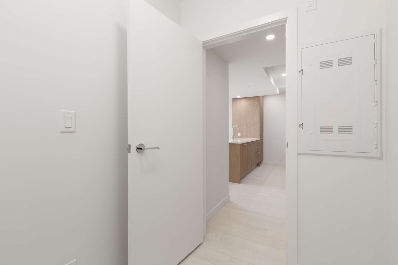 Open door leading to a bright room with wooden cabinetry and a visible sink. White walls and light flooring are visible.