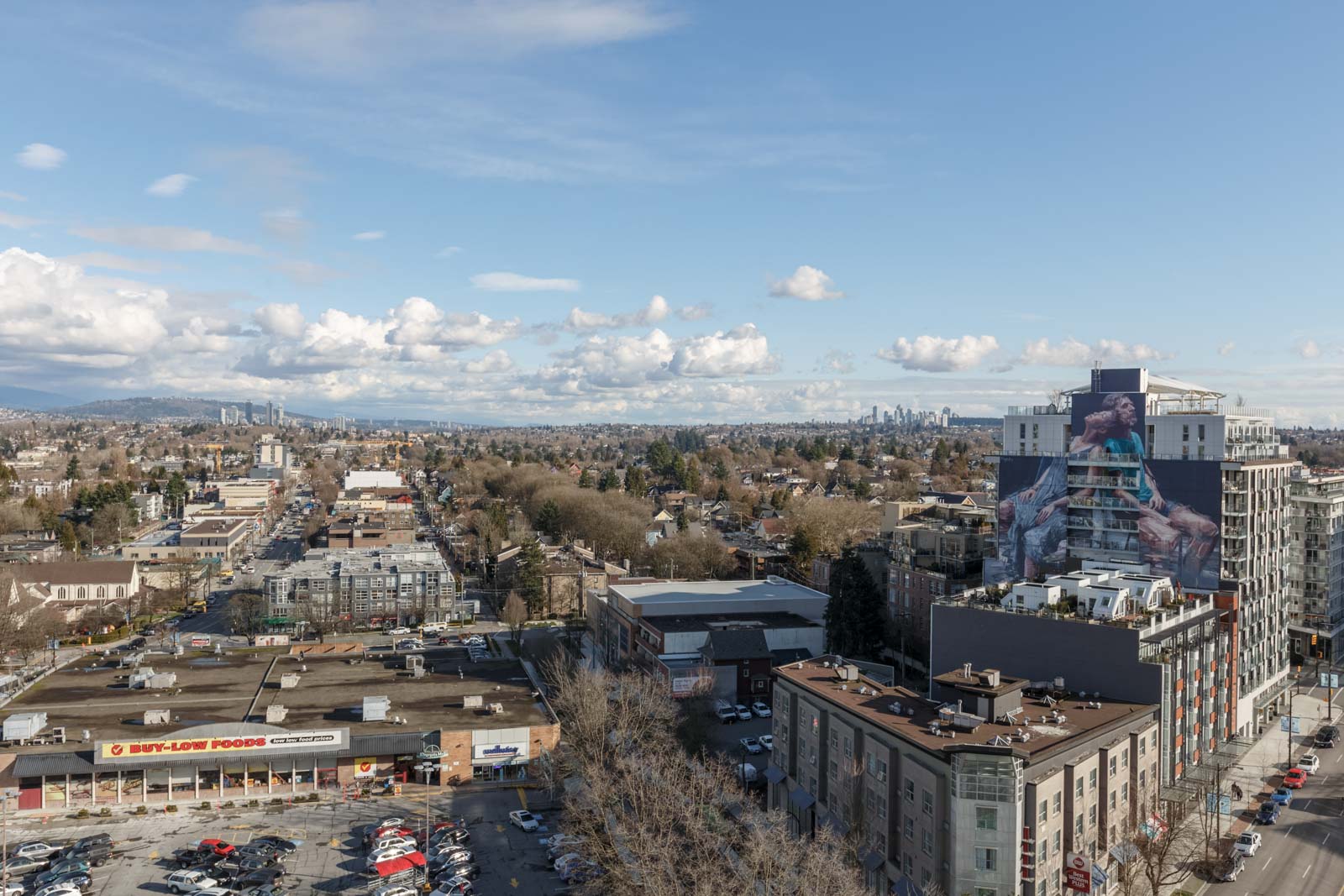 Aerial view of a cityscape with a grocery store parking lot in the foreground and distant city skyline under a cloudy blue sky.