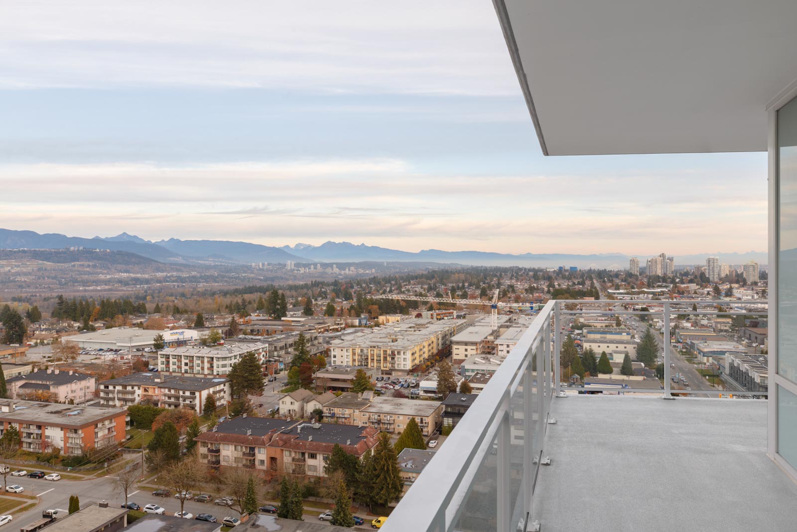 View from a high-rise balcony overlooking a suburban cityscape with buildings, streets, trees, and mountains in the distance under a cloudy sky.