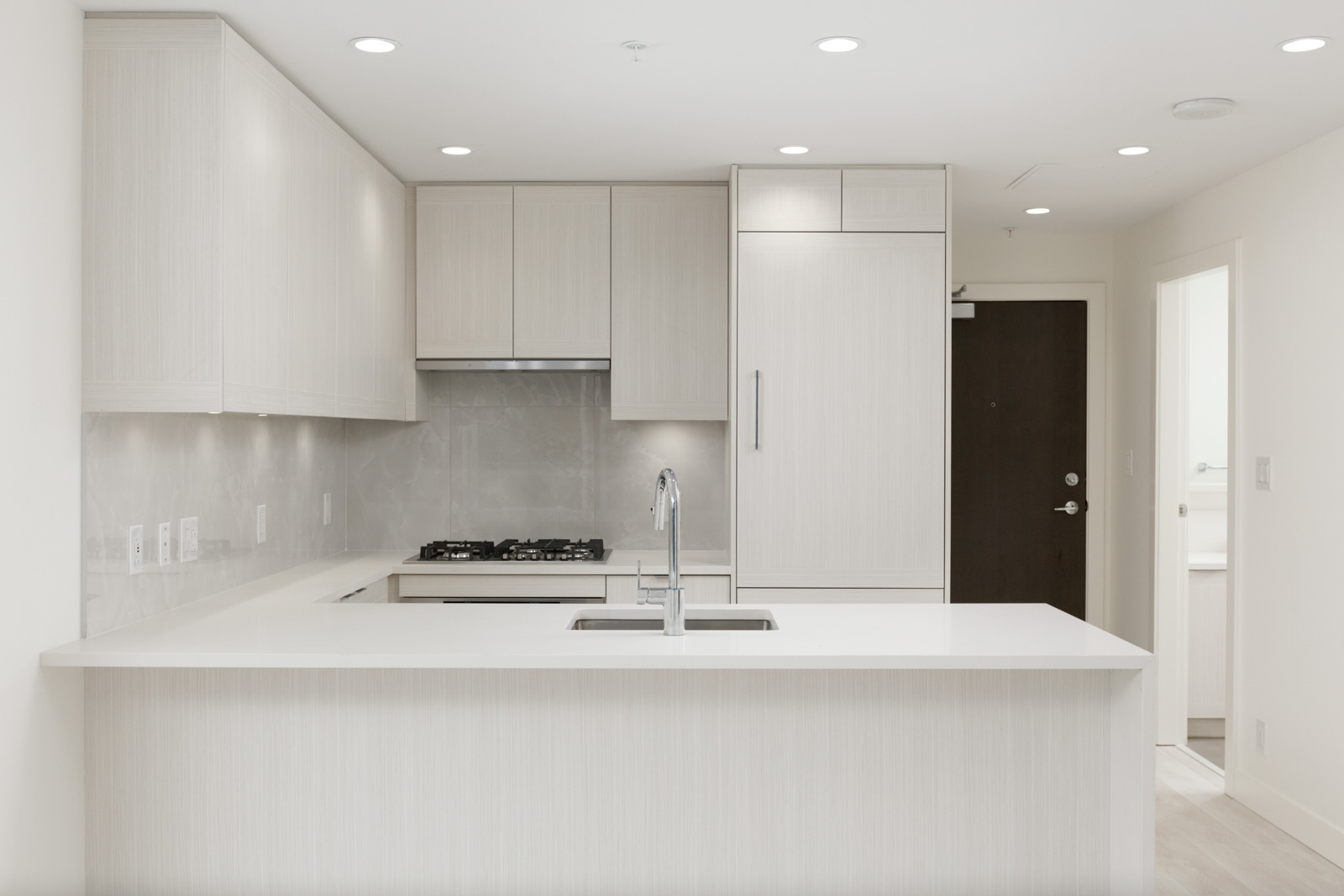 Modern kitchen with light-colored cabinets, built-in appliances, gas stove, under-cabinet lighting, and a large island with a sink; door and hallway visible in the background.