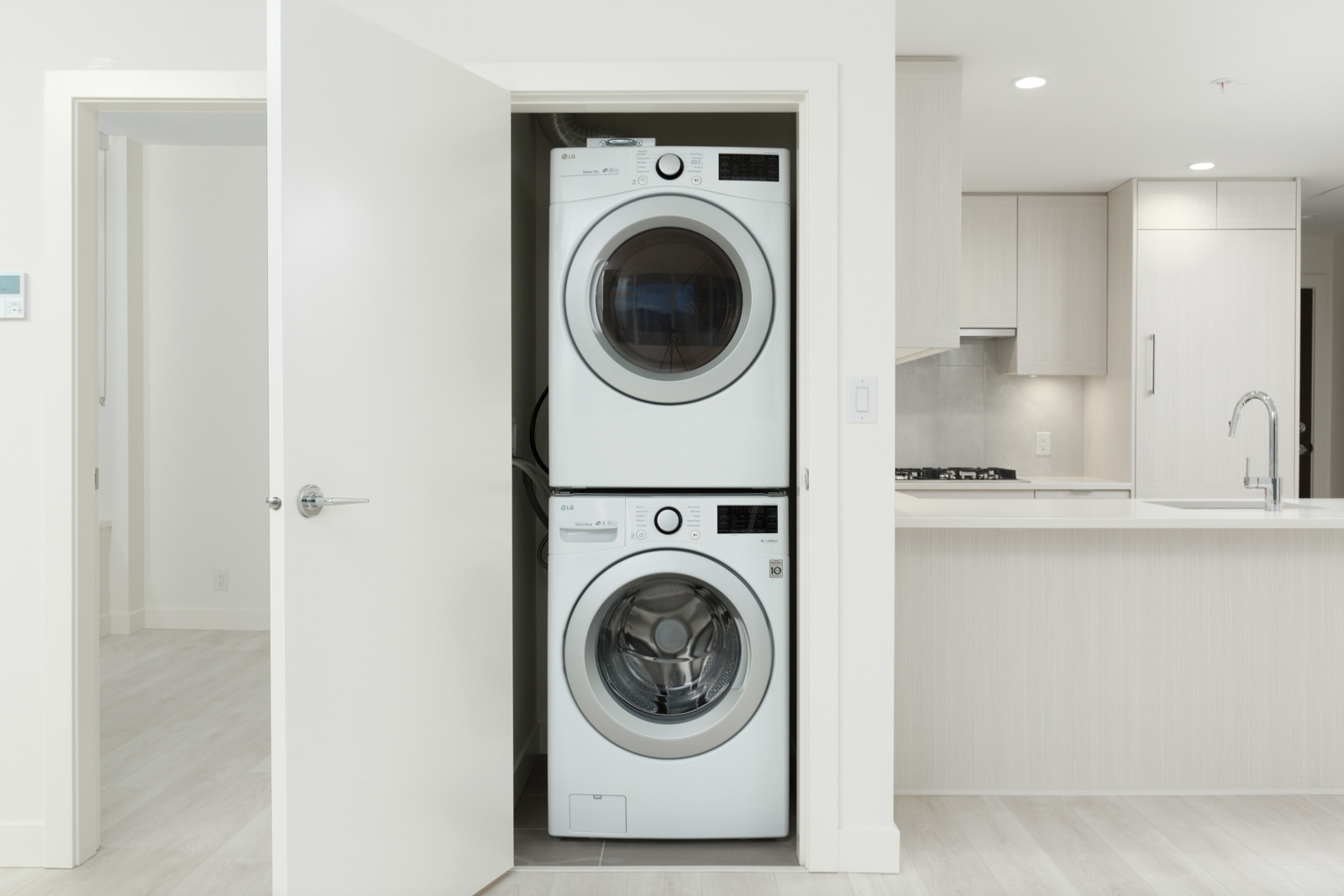 A stacked washer and dryer set is installed in a small closet next to a modern kitchen with white cabinetry and countertops.
