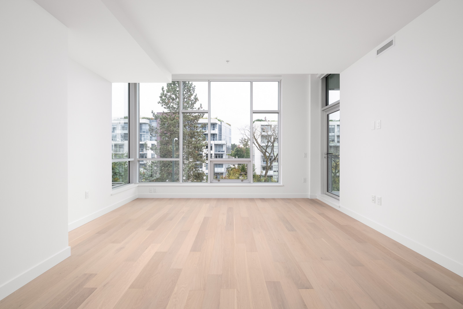 Empty room with light wood flooring, large windows, and white walls. Trees and buildings are visible outside.
