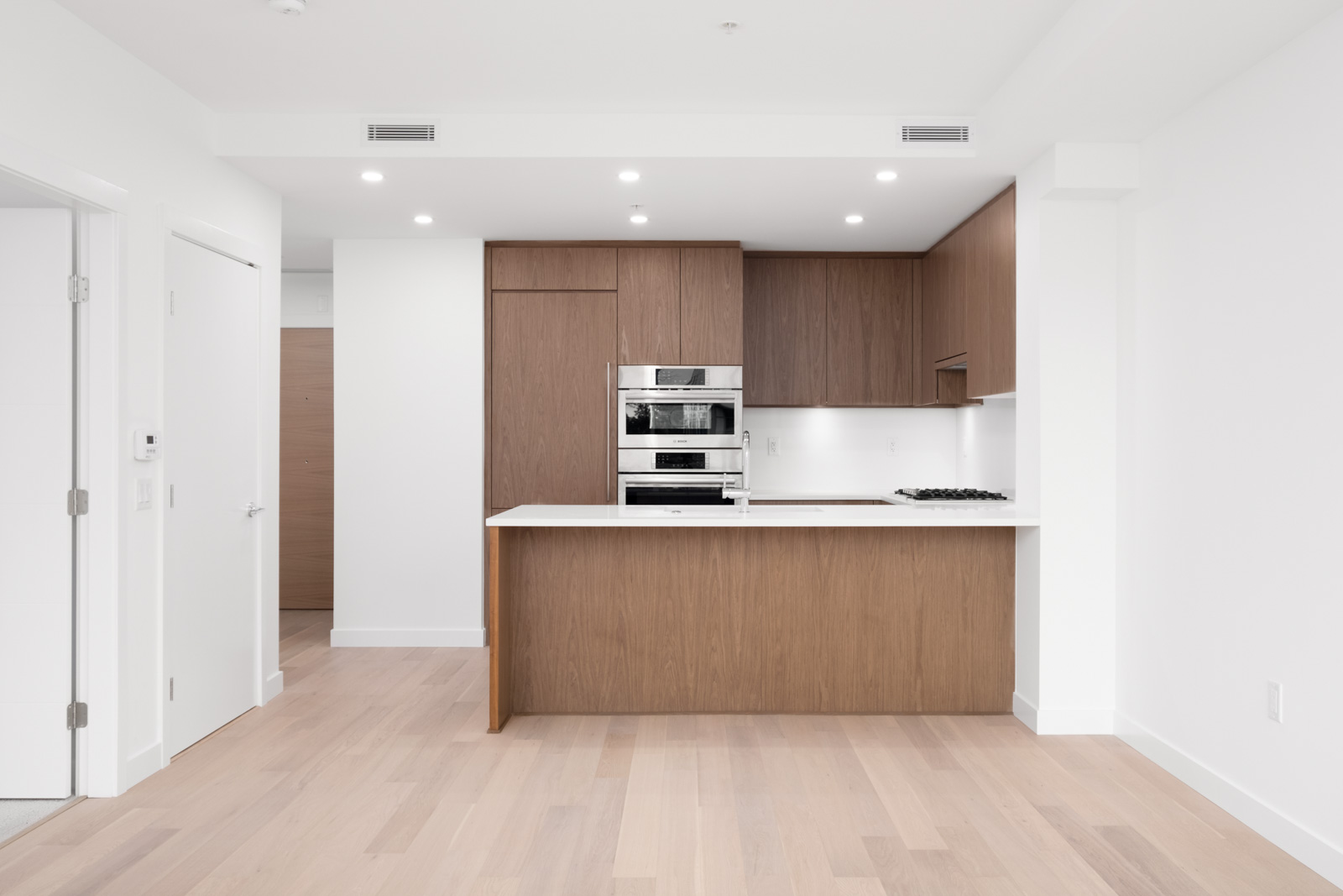 Modern kitchen with wooden cabinets, white countertops, built-in appliances, and light wood flooring. Minimalist design with recessed lighting and an open layout.