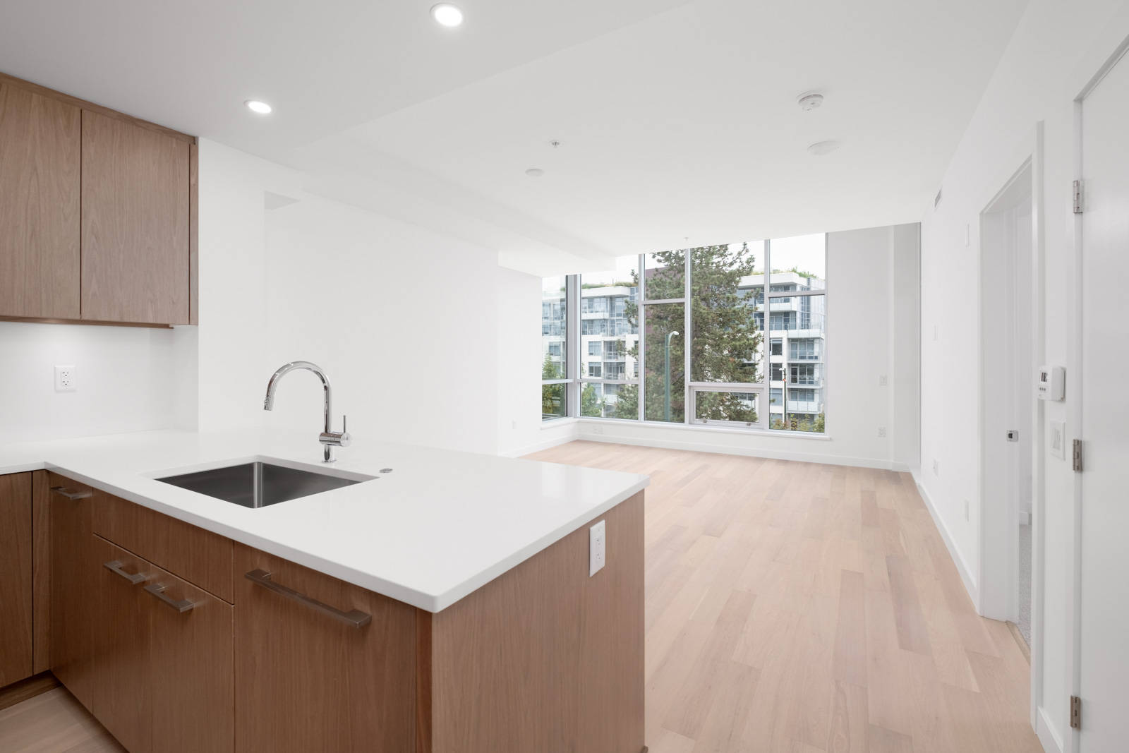 Modern, unfurnished apartment interior with wooden floors, large windows, and a kitchen featuring a white countertop and sink.