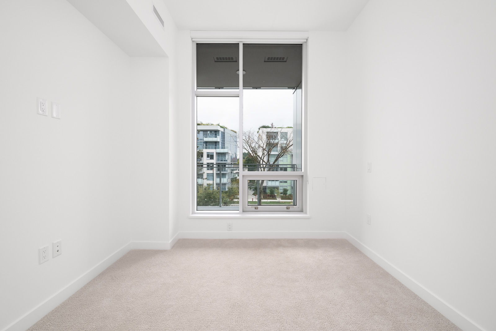 Empty room with beige carpet and large window overlooking modern buildings and trees outside. Walls are white, and there are electrical outlets on the left.