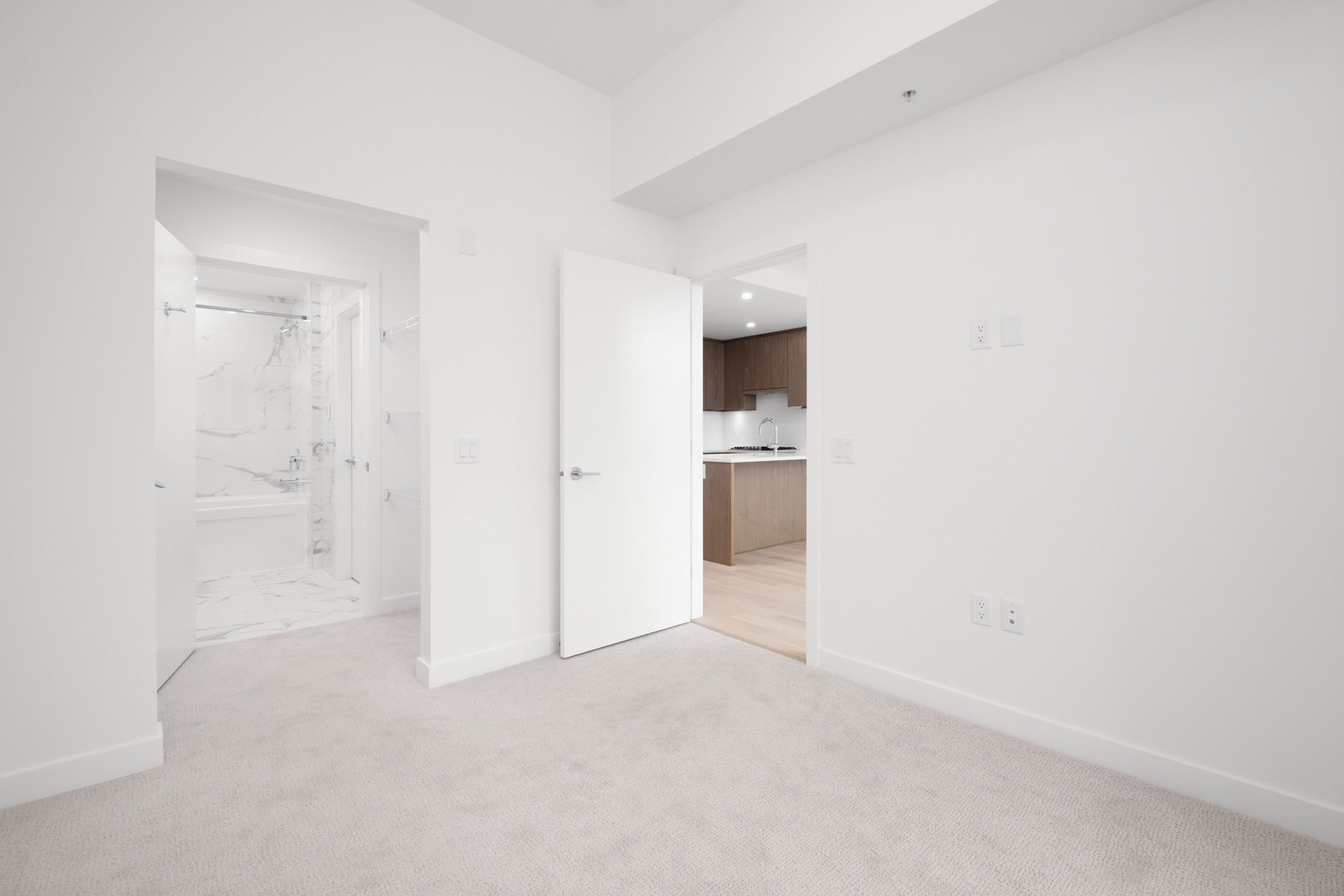 Minimalist room with white walls and carpet, featuring a door leading to a bathroom with marble tiles on the left, and a partially open door revealing a kitchen with wooden cabinets on the right.