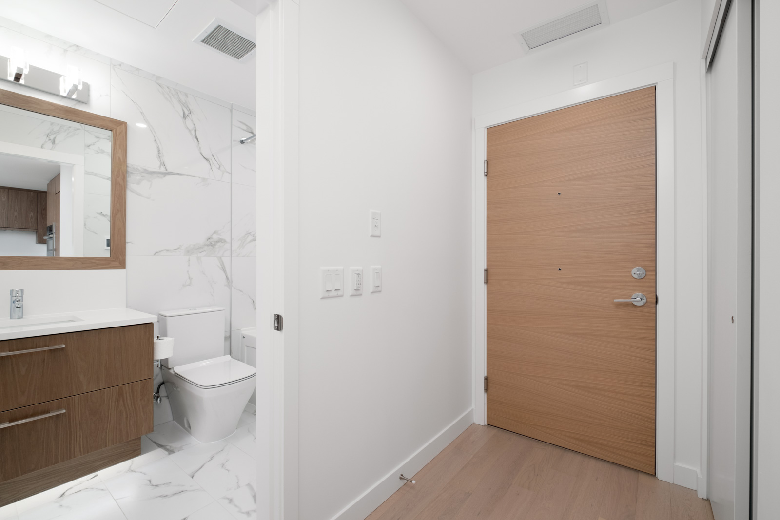 Modern bathroom with wooden cabinets, white countertops, and marble walls. Adjacent is a closed wooden door leading to a small hallway with wood flooring.