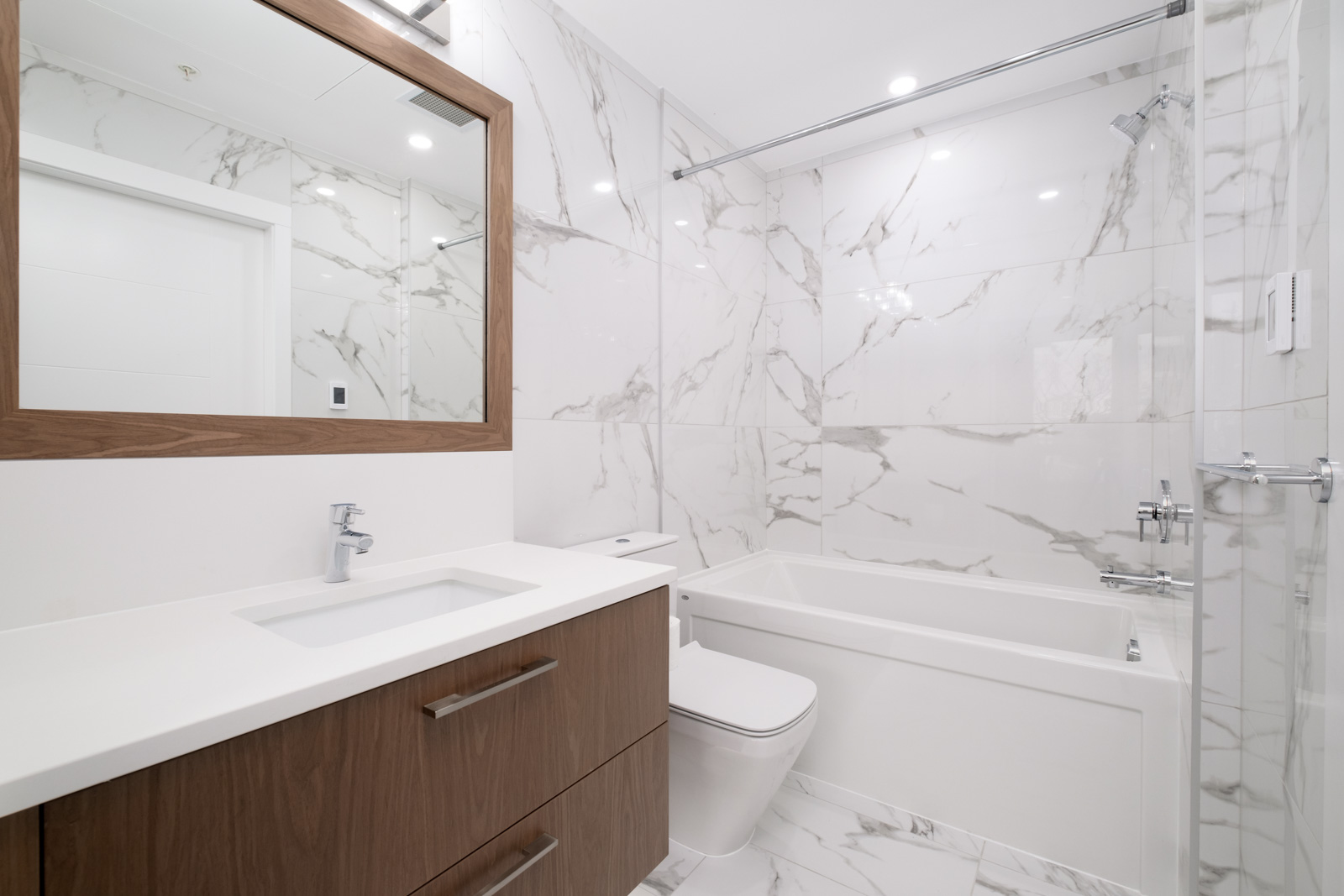 Modern bathroom with white marble walls, wooden vanity with a sink and mirror, toilet, and bathtub with a showerhead.