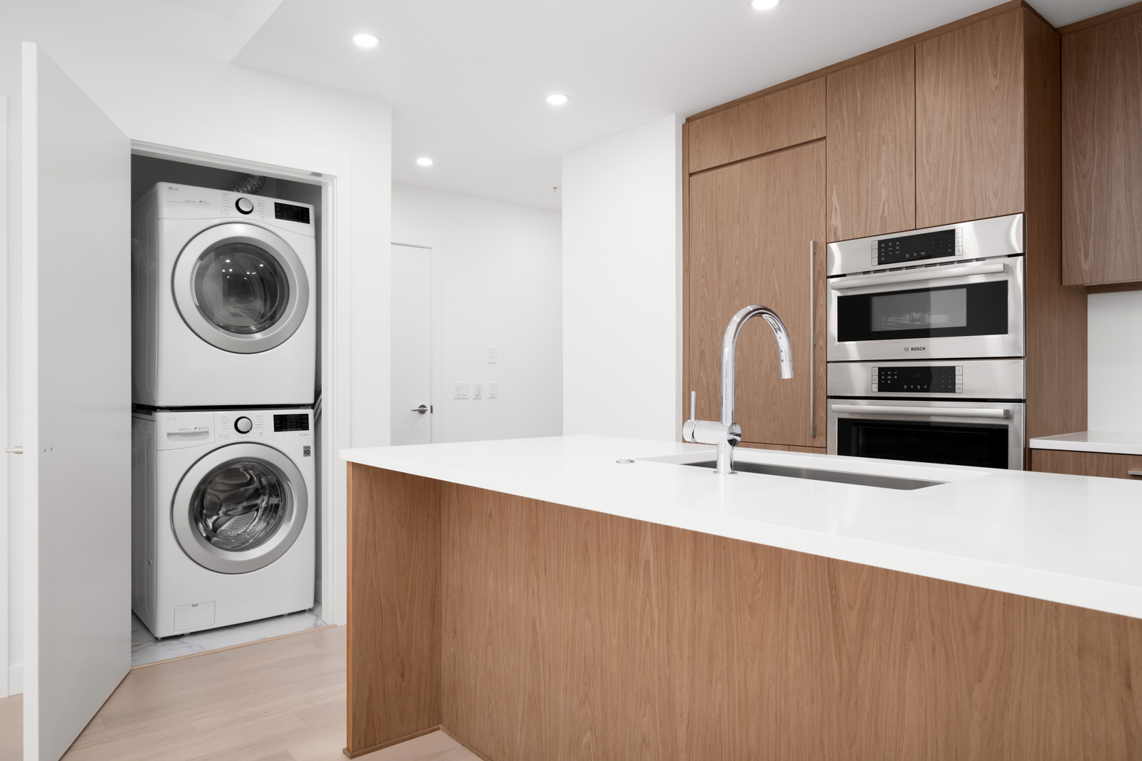 Modern kitchen with wooden cabinetry, white countertop island, built-in appliances, and a stacked washer-dryer unit in a closet.