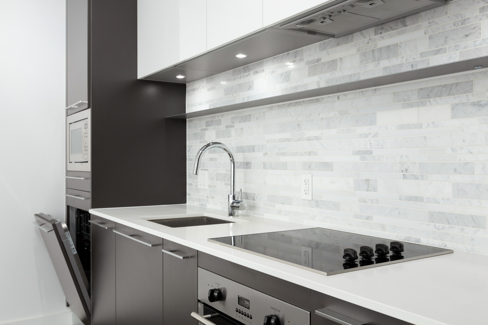 Modern kitchen with white countertops, electric stove, built-in oven, microwave, stainless steel sink, and grey and white tile backsplash.