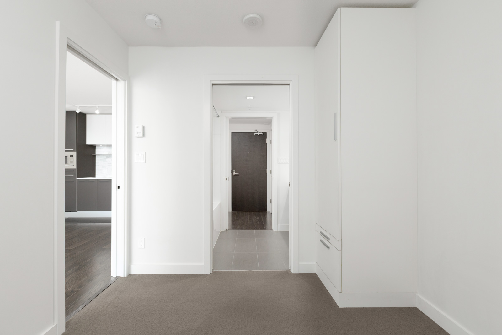 A hallway in a modern apartment with white walls, beige carpet, and built-in storage; doorways lead to a kitchen and an entryway with a dark door.