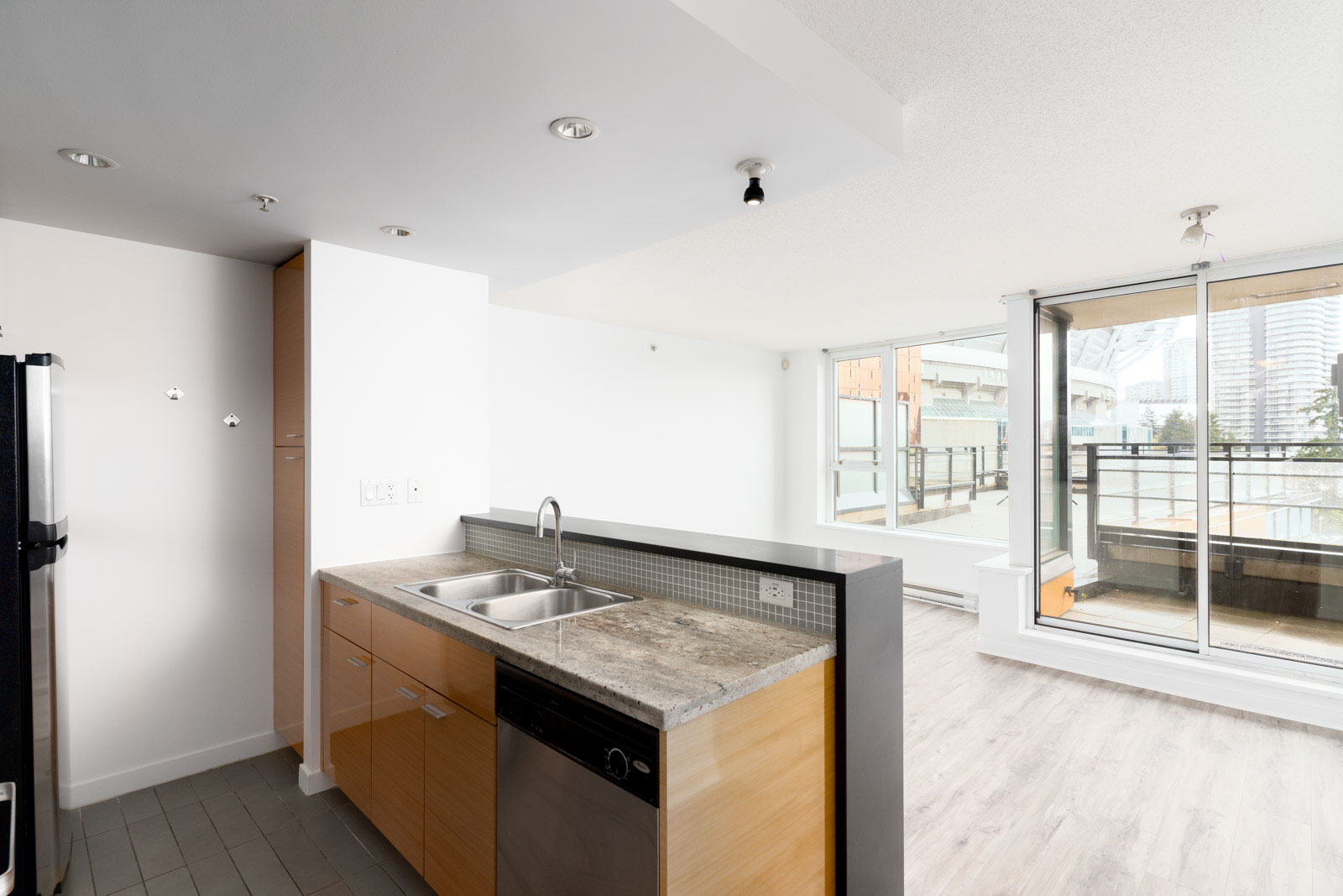 Unfurnished apartment interior with an open kitchen, wooden cabinets, sink, and a living area with large windows and balcony access.
