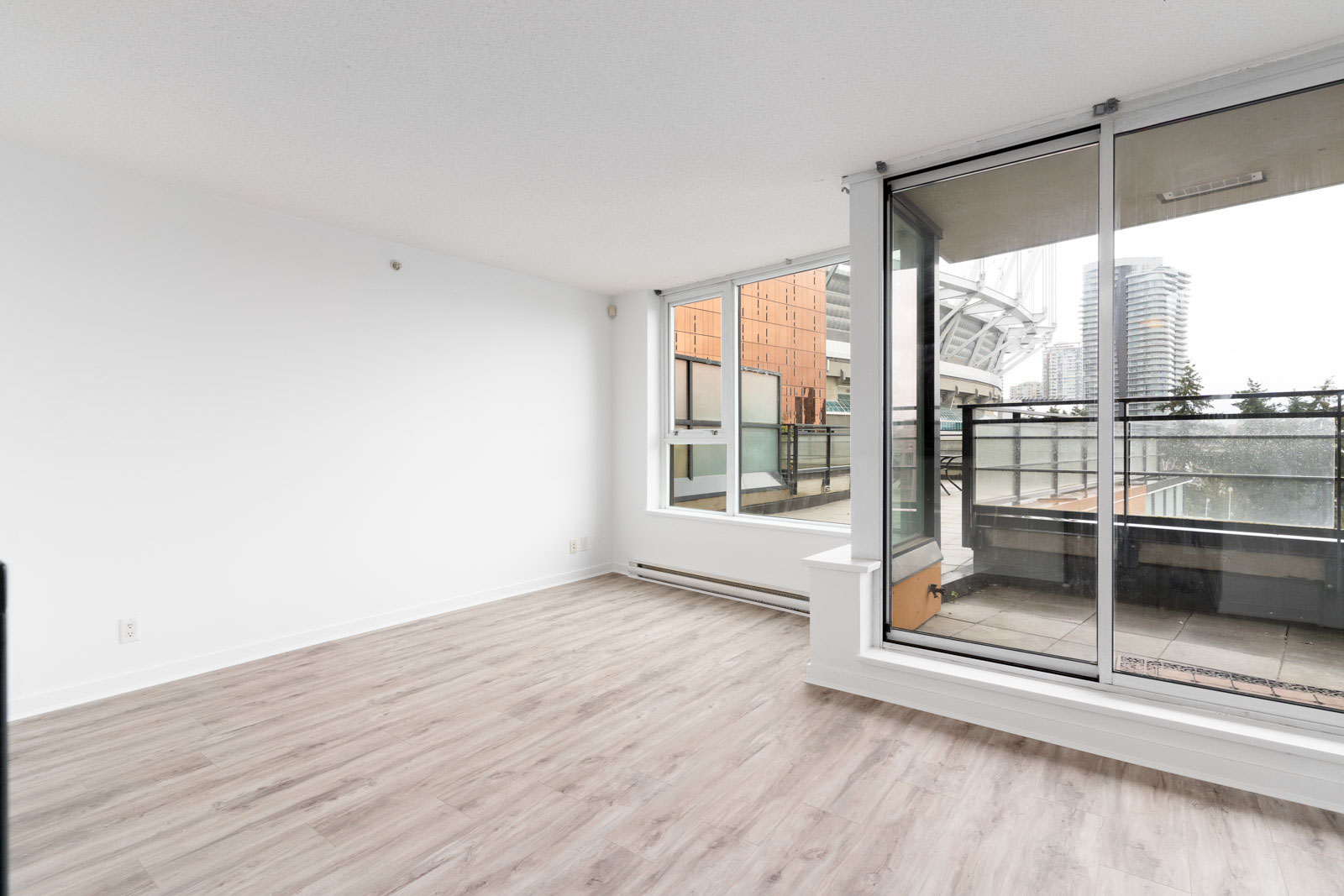 Empty room with light wood flooring, white walls, and large glass doors leading to a balcony. City buildings visible through the windows.