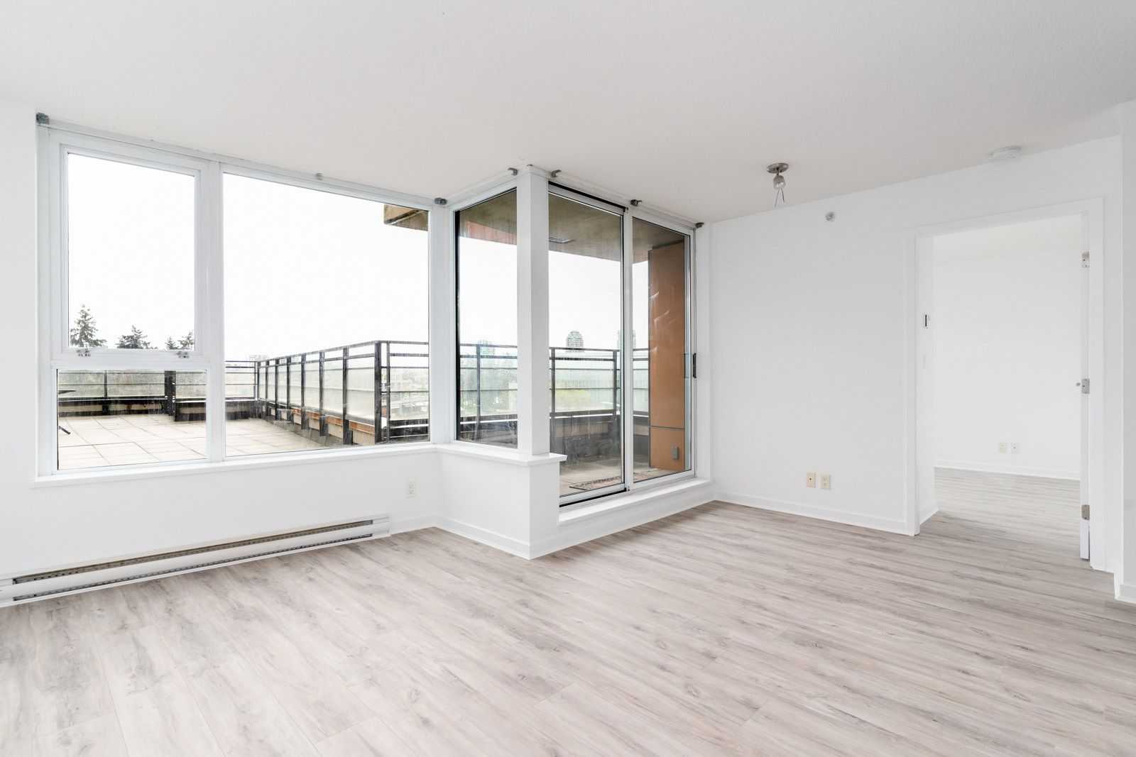Empty room with light wood flooring, large windows, and a door leading to a balcony. Walls are white, and there is a ceiling light fixture. Doorway opens to another room.