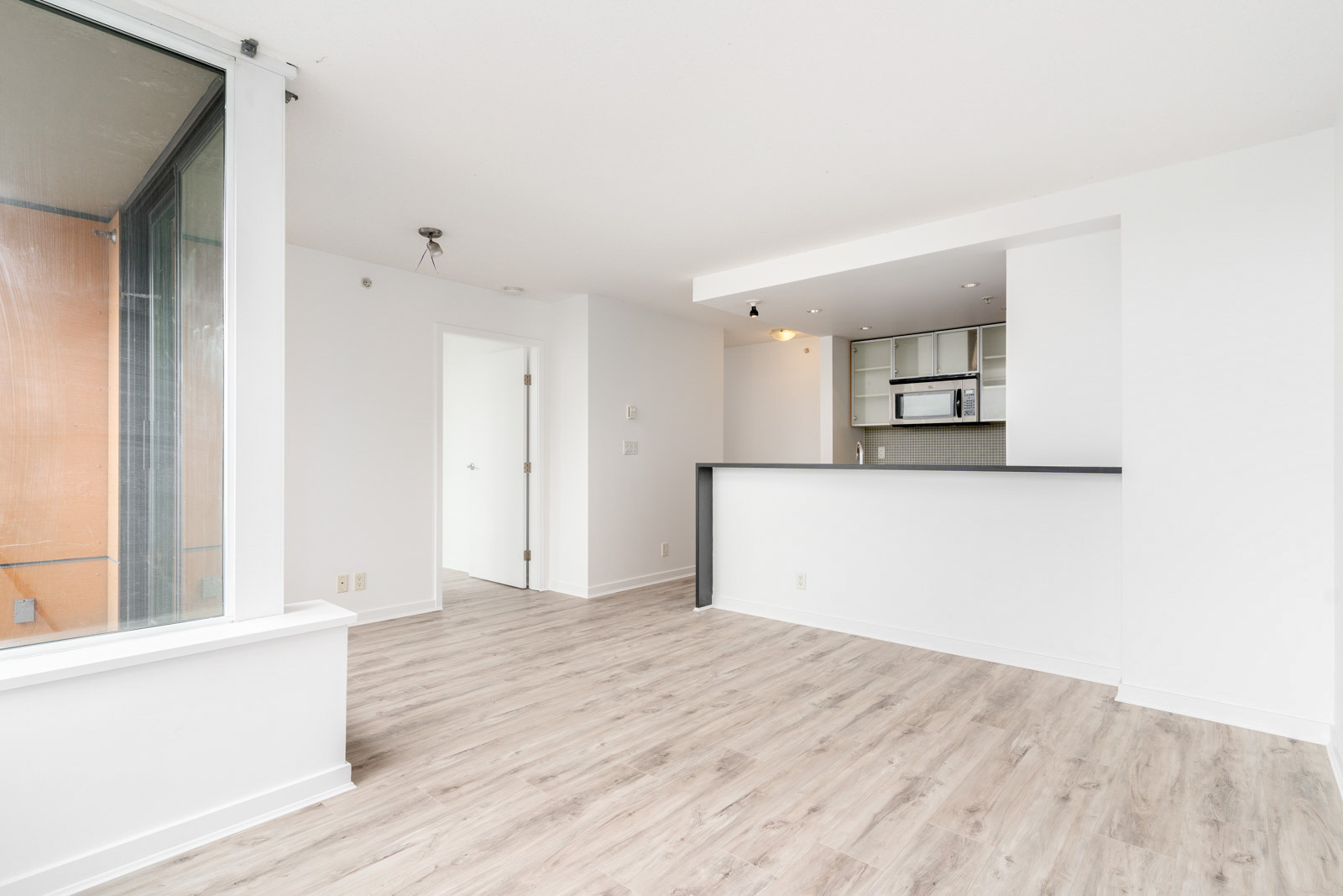 Spacious, unfurnished apartment with light wood flooring, open kitchen, large window, and white walls.