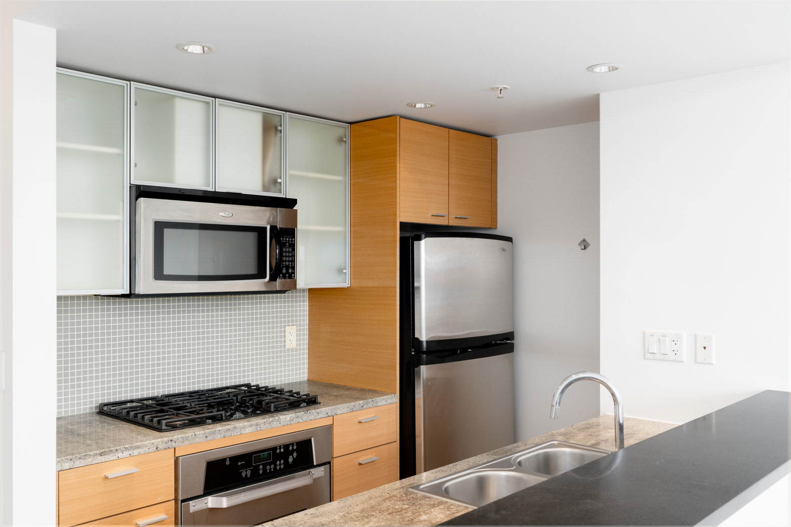 Modern kitchen with stainless steel appliances, including a microwave and refrigerator, wood cabinets, gas stove, and a double sink. Gray tile backsplash and light countertops are visible.