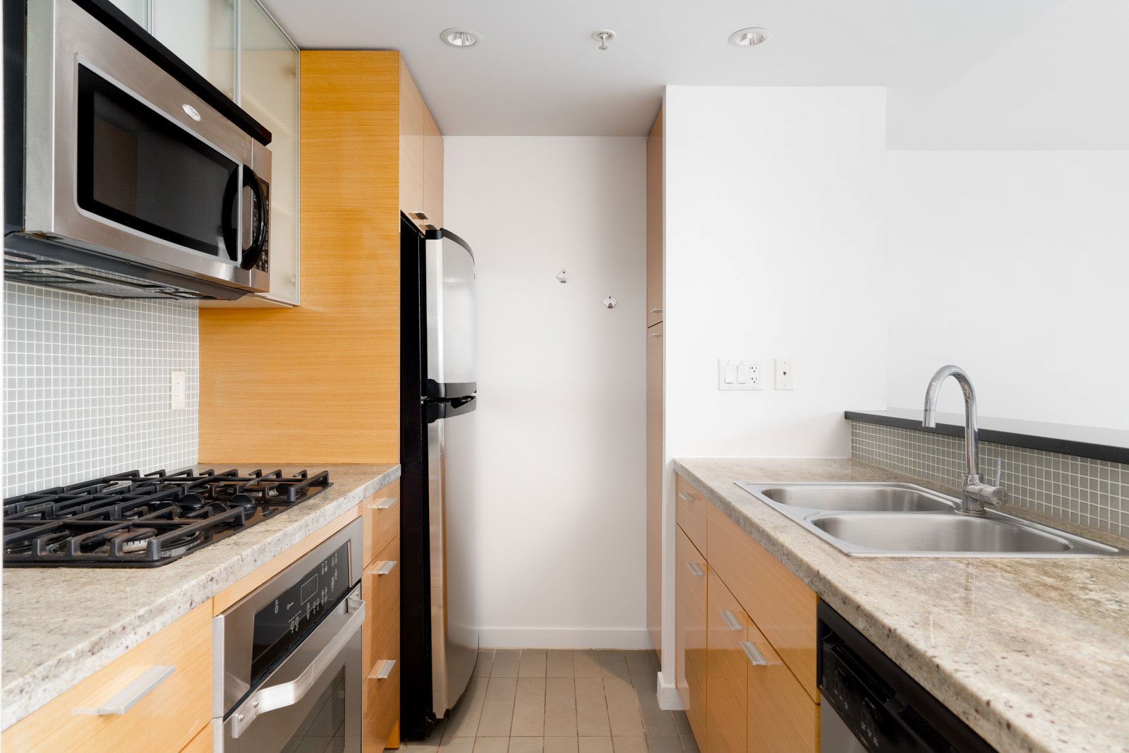 Modern kitchen with stainless steel appliances, including a microwave, oven, and refrigerator. Light wood cabinets, stone countertops, double sink, and a tiled backsplash.