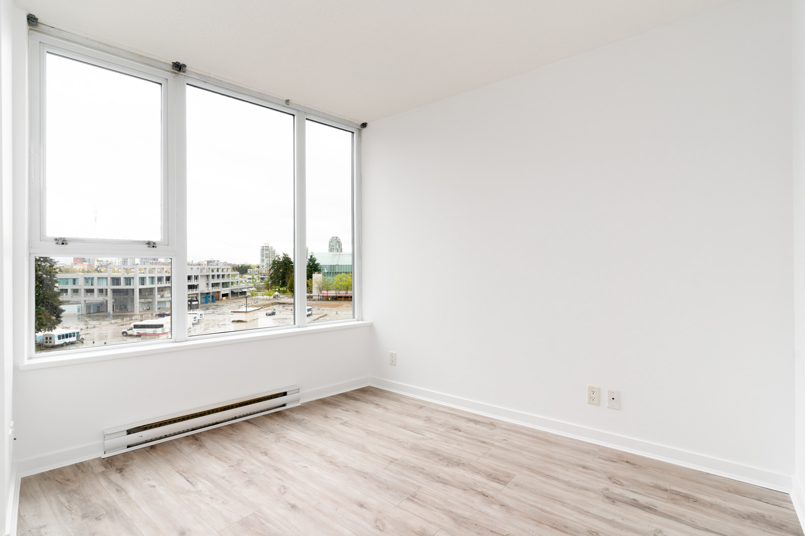 An empty room with white walls, large windows, and light wood flooring. Urban view outside.