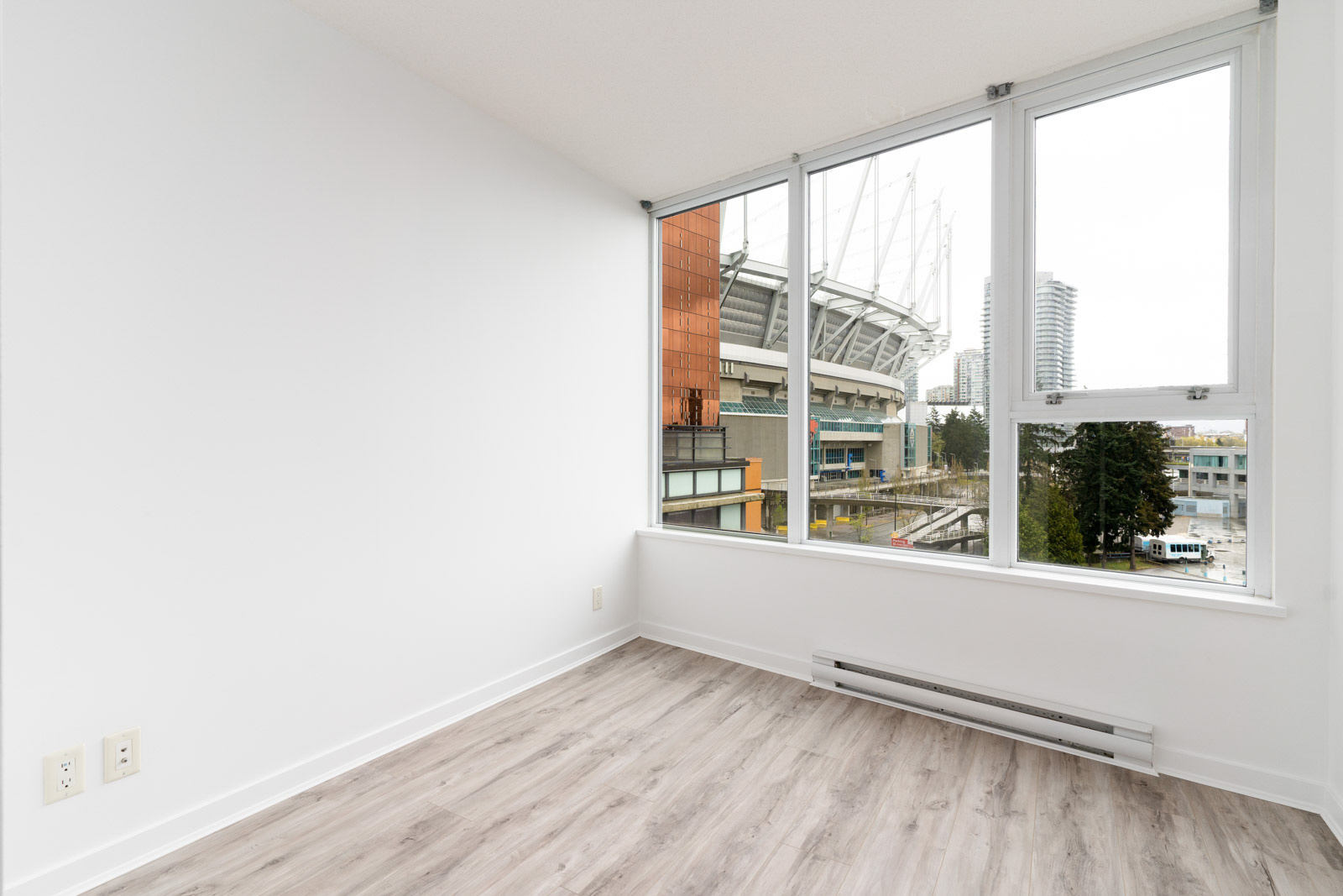 Empty room with light wood flooring and large windows showing a stadium and cityscape outside.