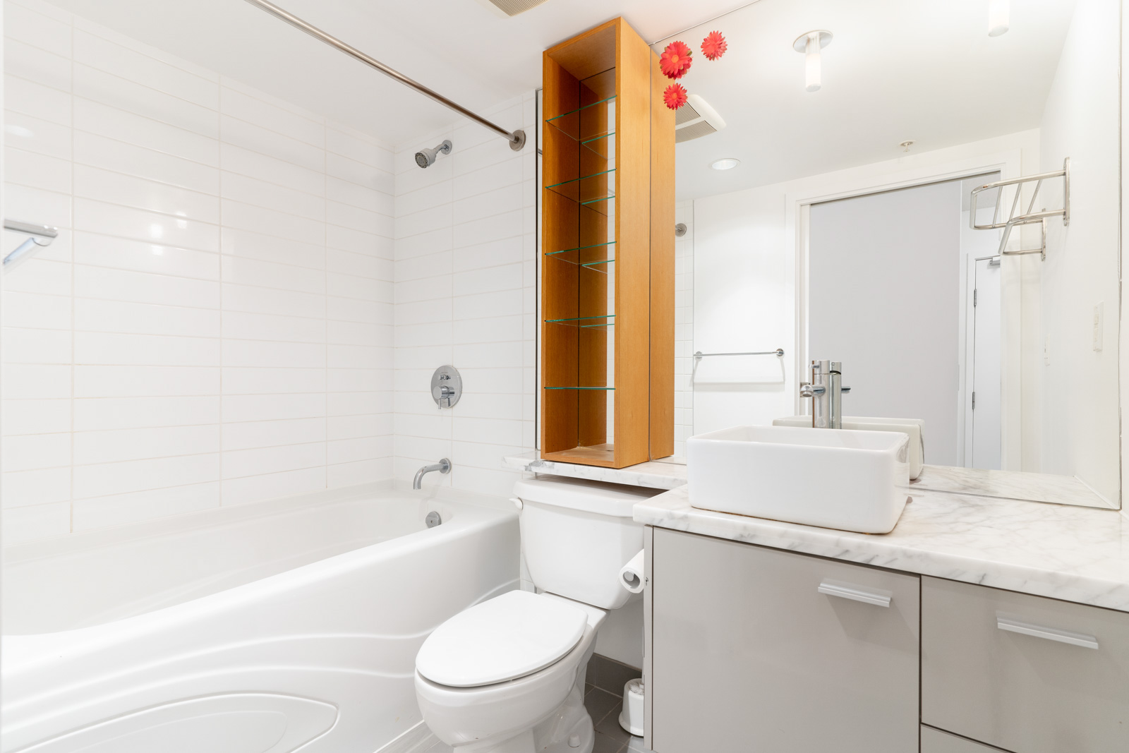Modern bathroom with a white bathtub, sink, and toilet. Wooden shelves above the toilet, marble countertop, and white tiled walls.