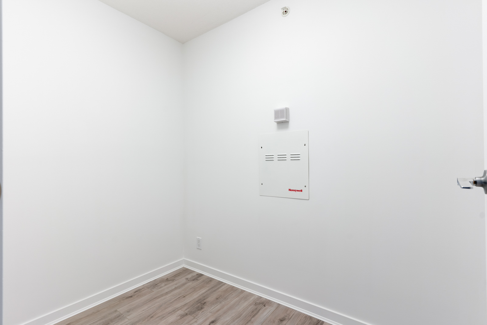 A small empty white room with light wood flooring, a wall vent, and a visible electrical panel.