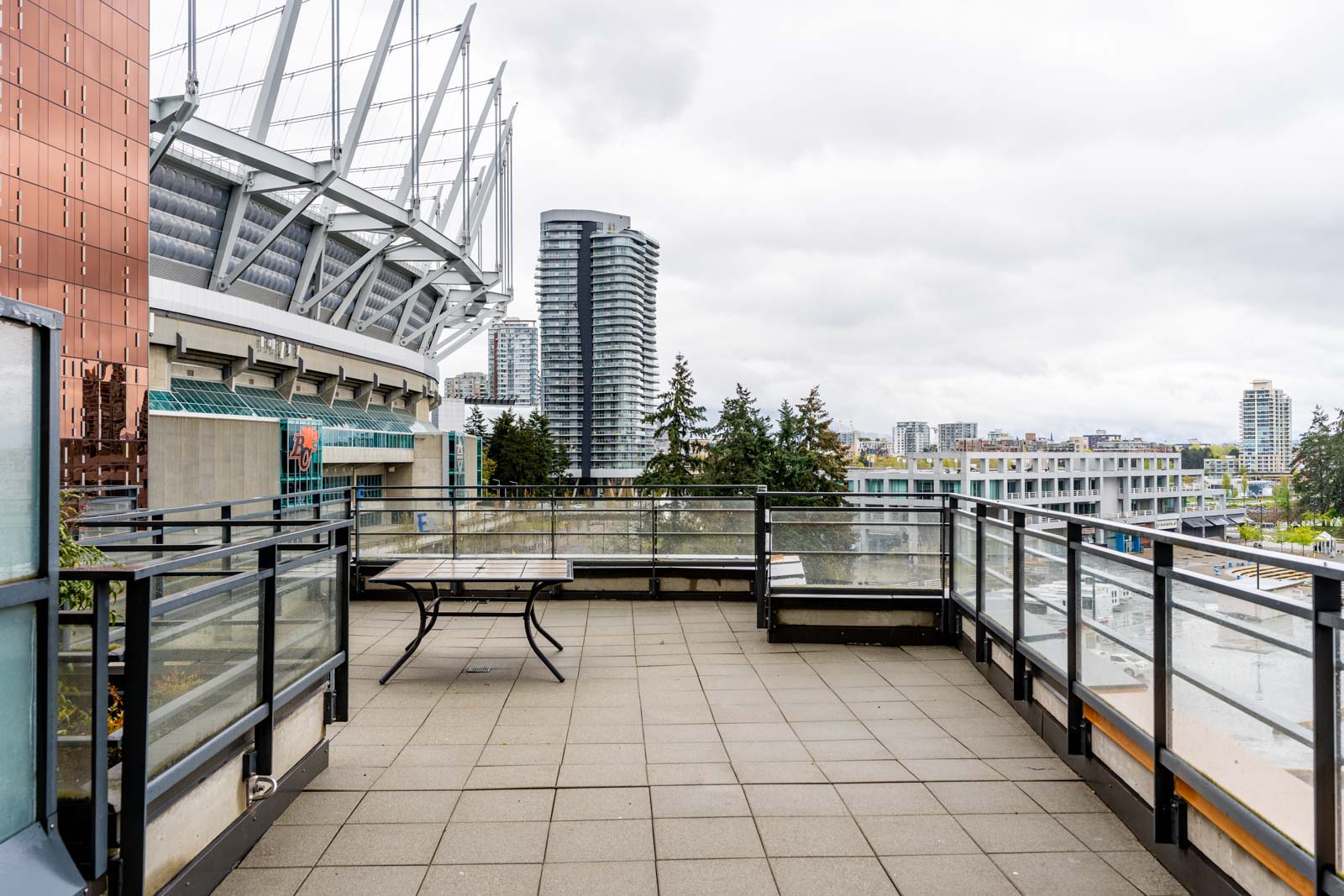Rooftop terrace with seating, metal railing, and city skyline view. Nearby are tall buildings and a large stadium structure under a cloudy sky.